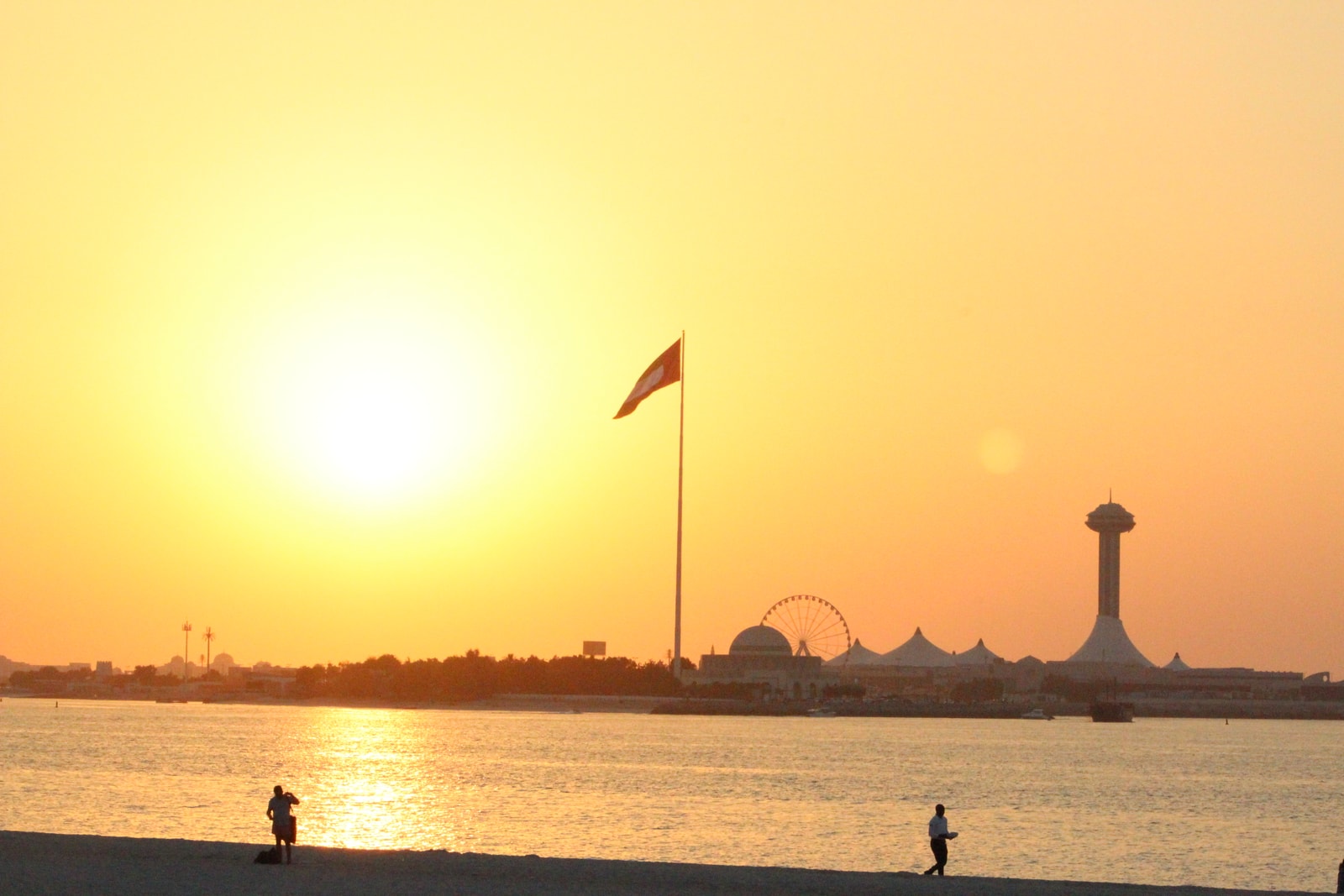 The sun sets over Corniche Beach in Abu Dhabi, casting a warm orange glow on the water and silhouetting buildings and a flag pole.