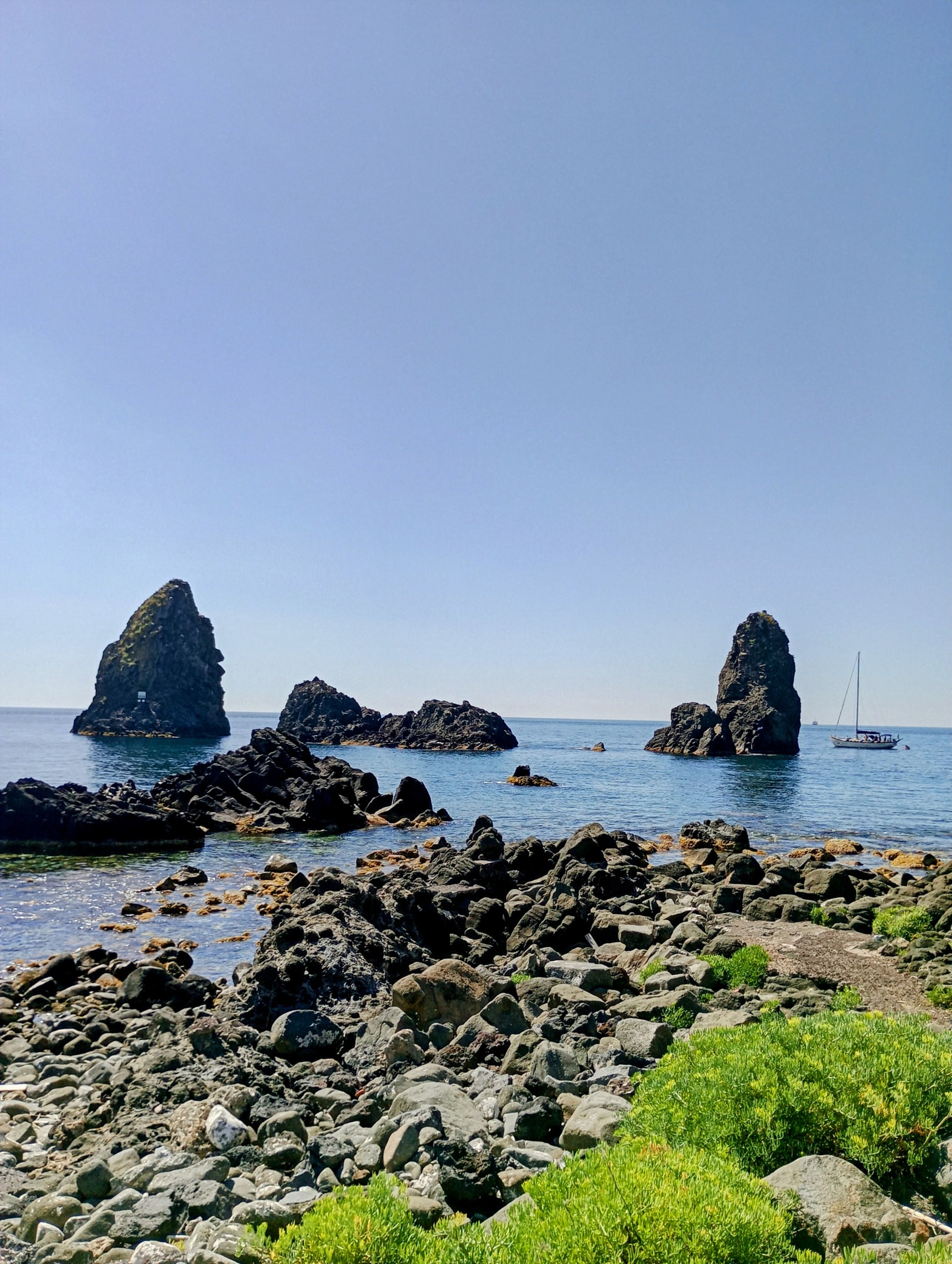 The basalt stacks of Aci Trezza in brilliant daylight, framed by sharp volcanic shoreline stones and a white sailboat on the horizon.