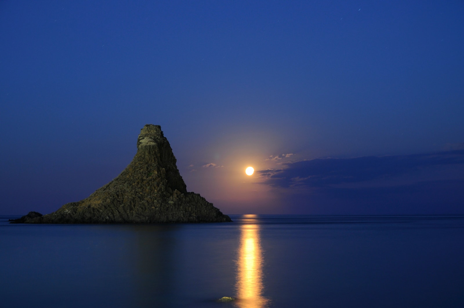 Atmospheric night shot showing a full moon rising out of the Ionian Sea next to the basalt stacks of Aci Trezza.