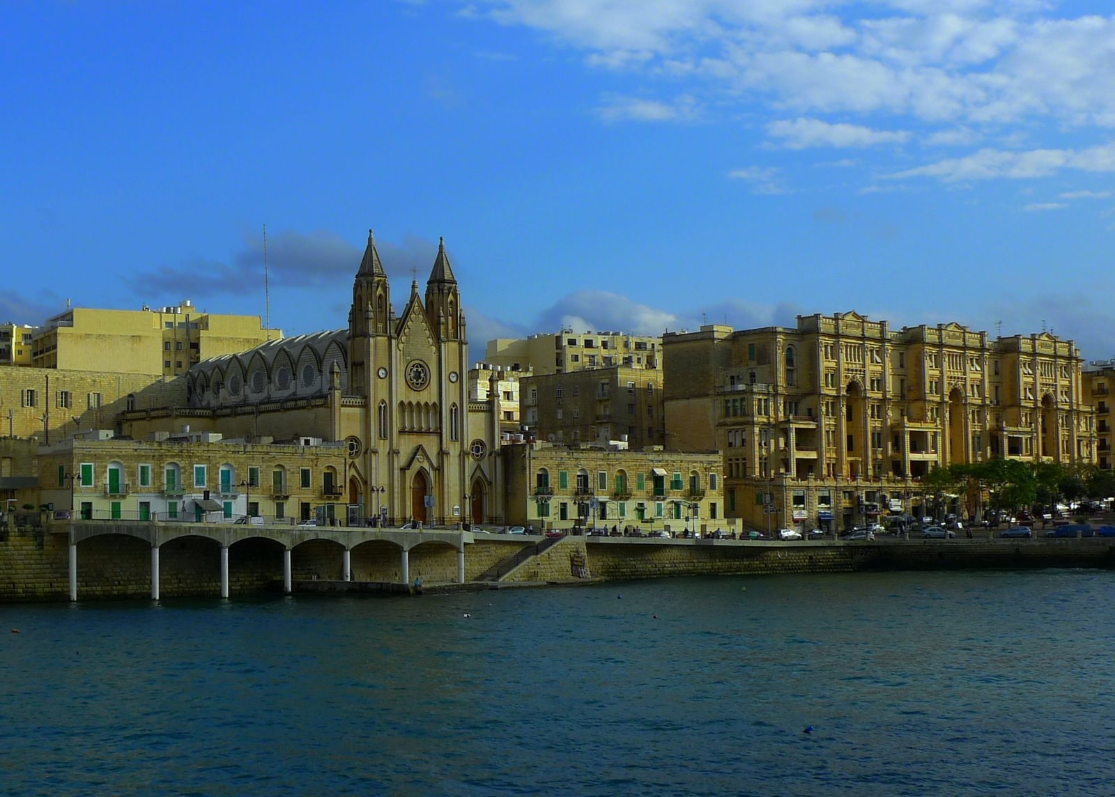 A view across the water of Balluta Bay in St. Julian's, Malta, framed by elegant Art Nouveau buildings and the striking Neo-Gothic Carmelite Parish Church.
