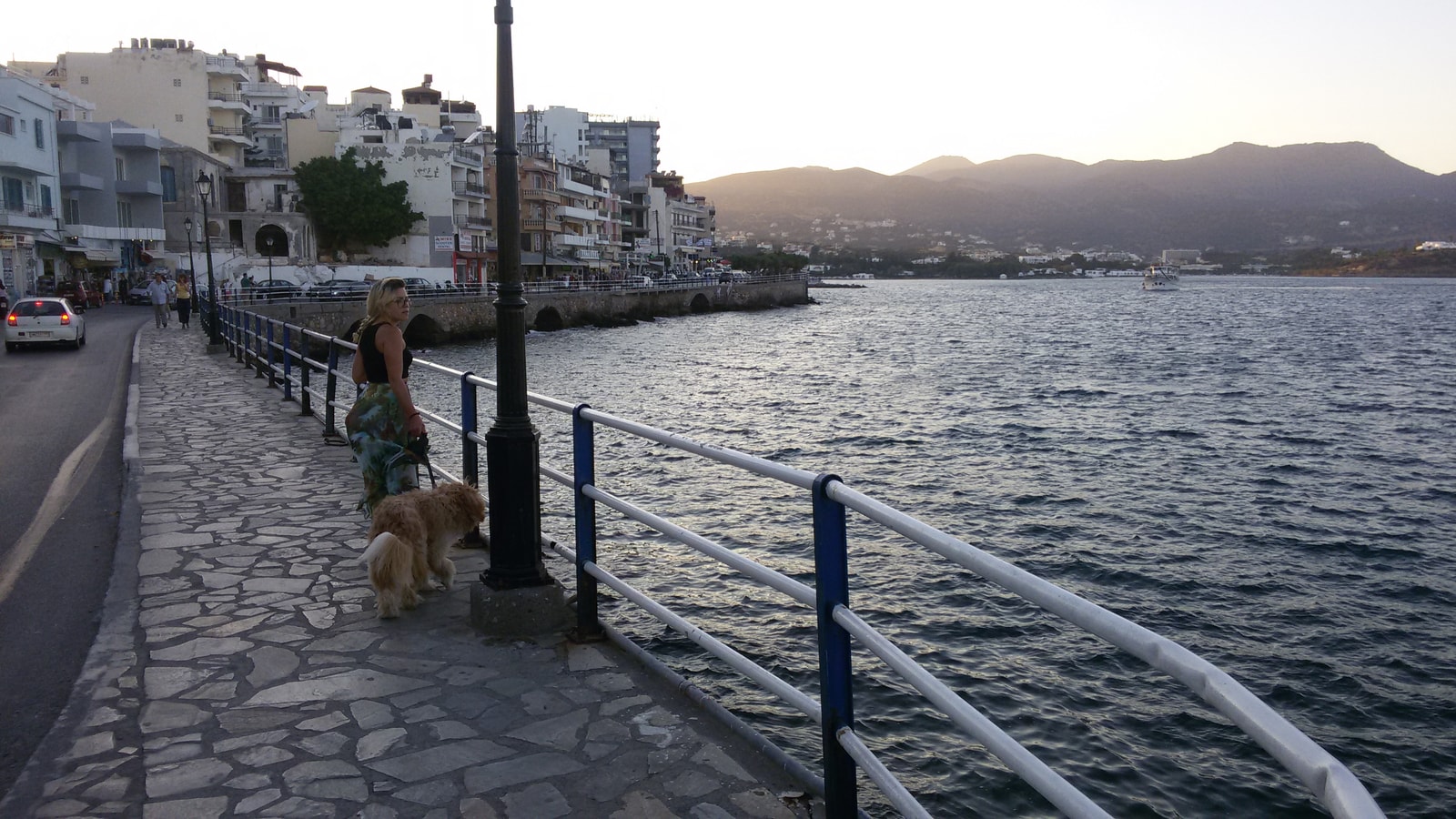 A paved coastal promenade in Agios Nikolaos with railings, with the blue sea and distant mountains in view.