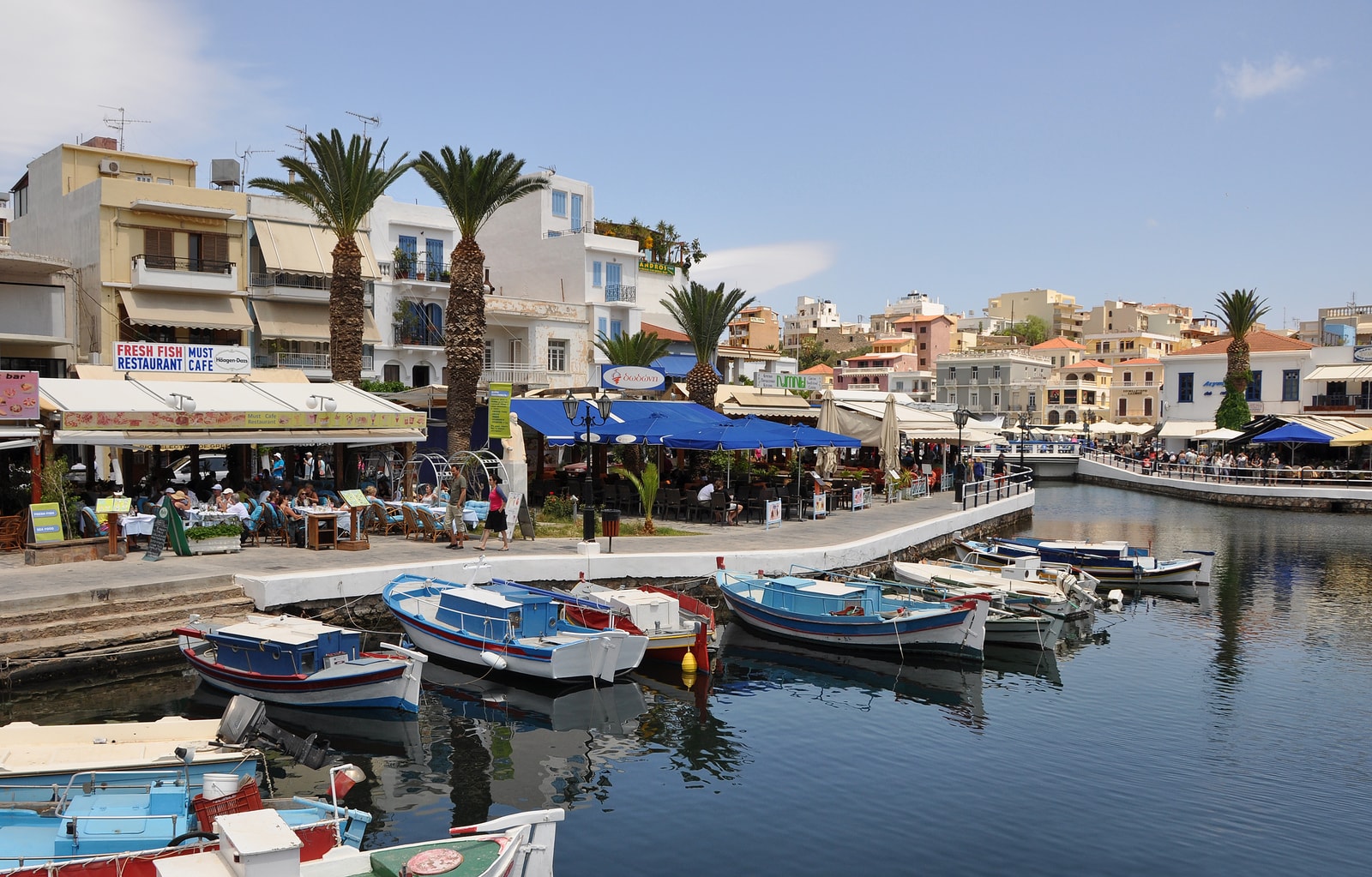 A sunny harbor scene in Agios Nikolaos with boats, palm trees, and waterfront cafes with umbrellas.