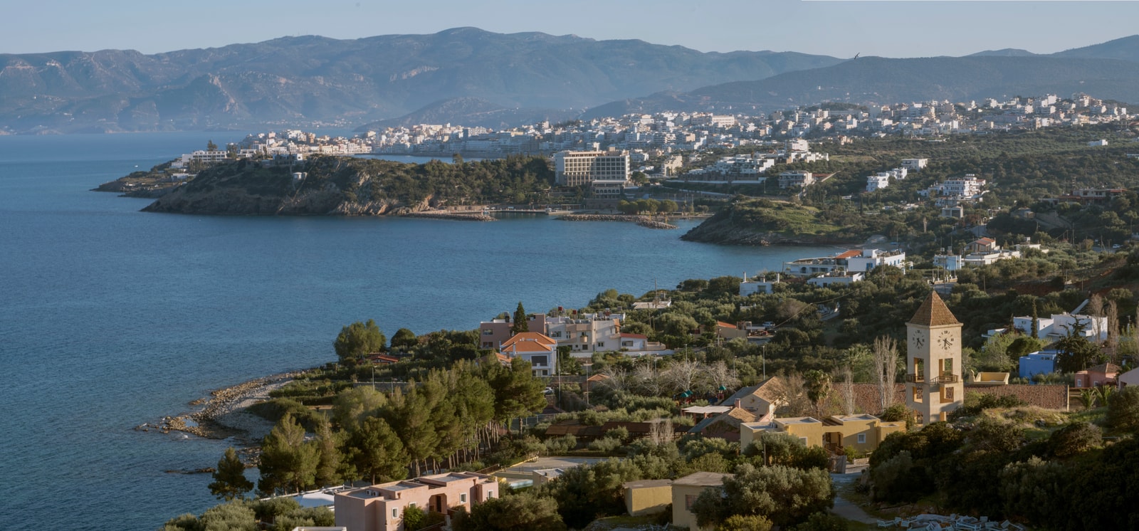 A wide panoramic view of Agios Nikolaos, showing the town spreading along the coastline with a bay and a mountainous background.
