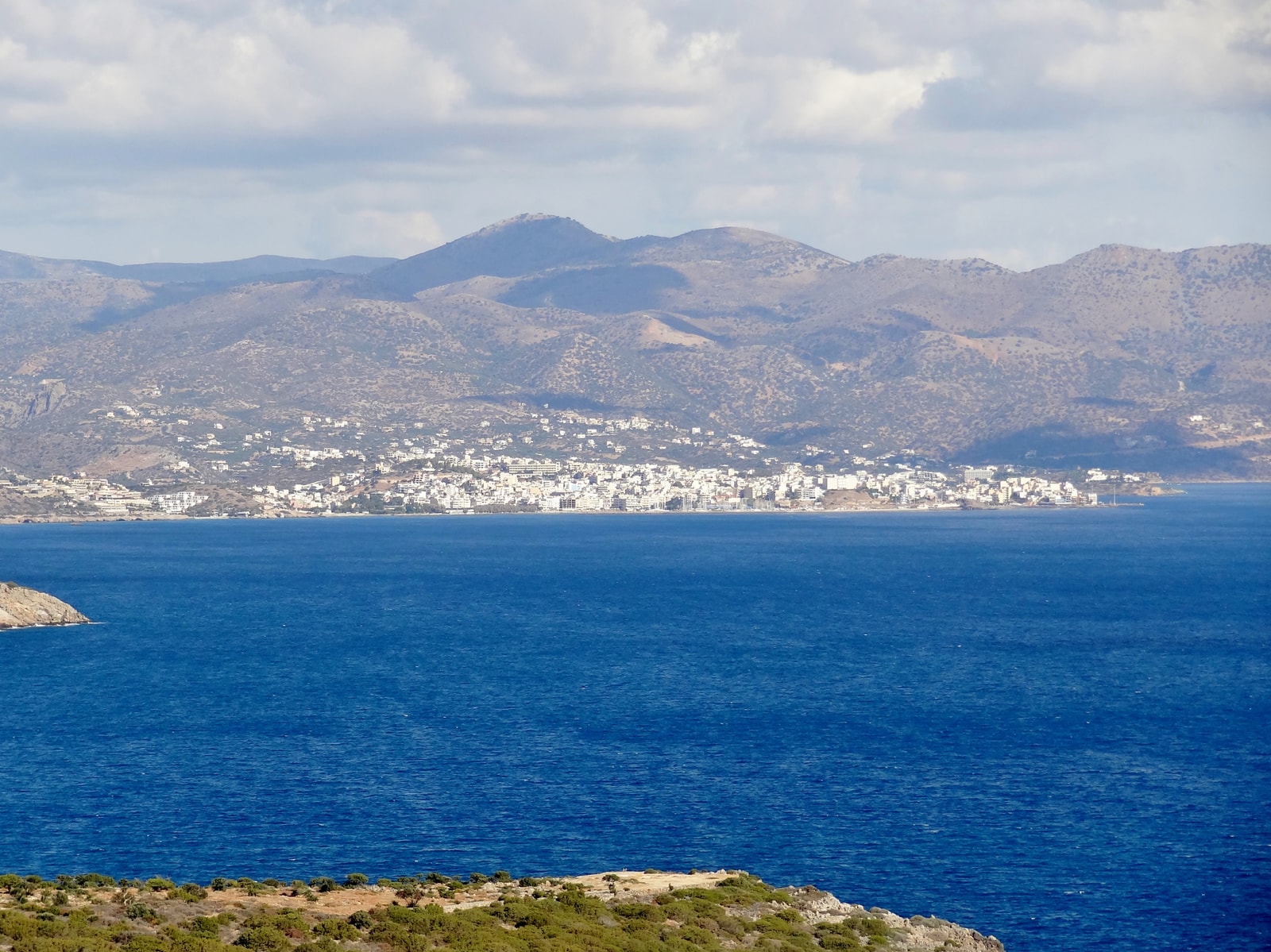 A wide view of Agios Nikolaos from across the sea, with a rocky foreground and mountains behind the town.