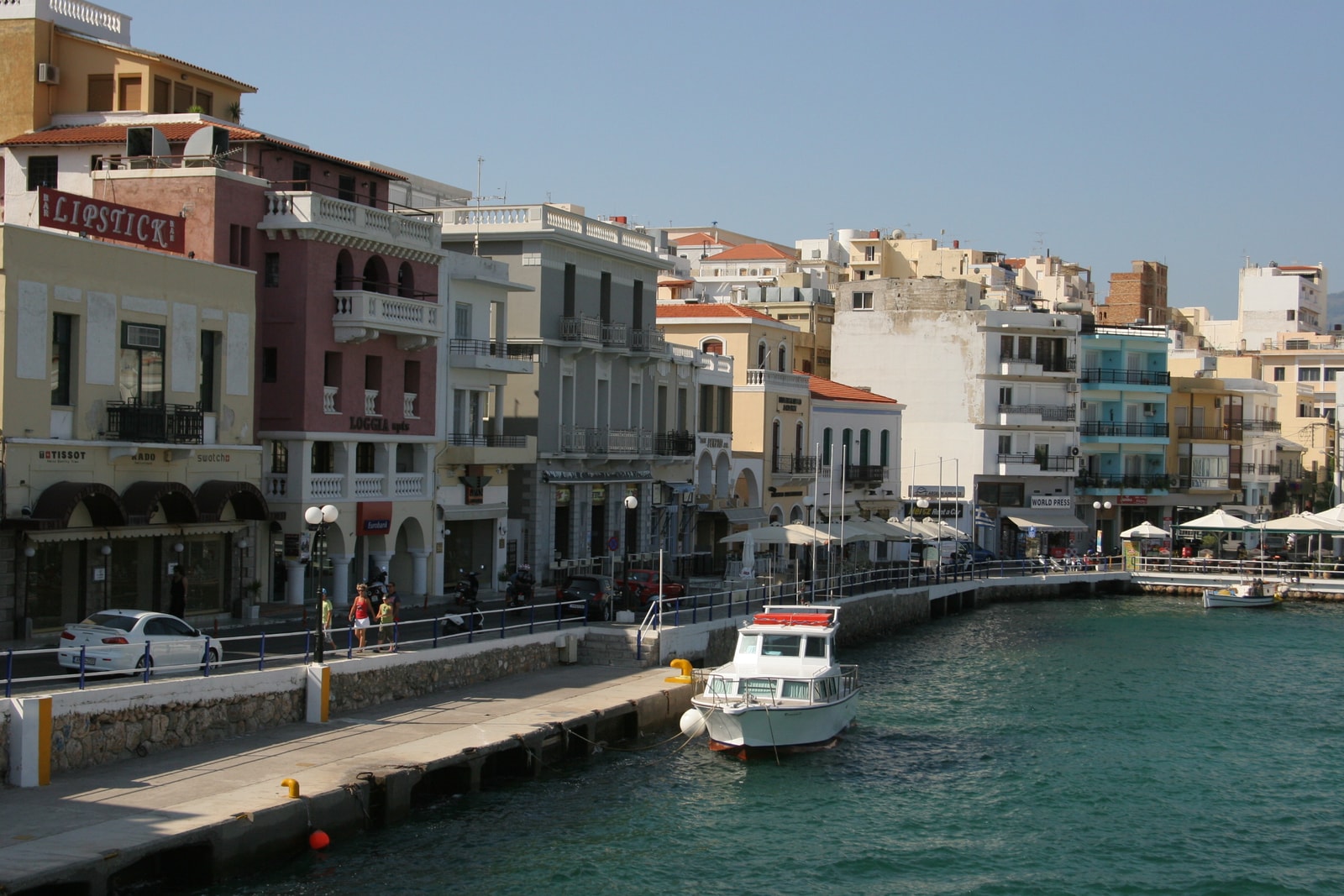 A passenger boat navigates a channel lined with waterfront cafes and quays in Agios Nikolaos.
