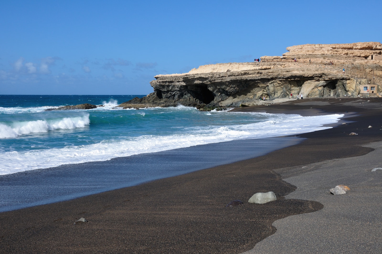 White waves crash against a dramatic shoreline of deep black sand, framed by towering sedimentary cliffs.