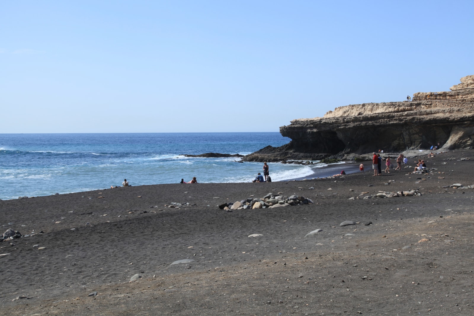 Massive limestone sea cliffs contain the dark openings of the Ajuy caves, with small figures of people on the path demonstrating the scale.