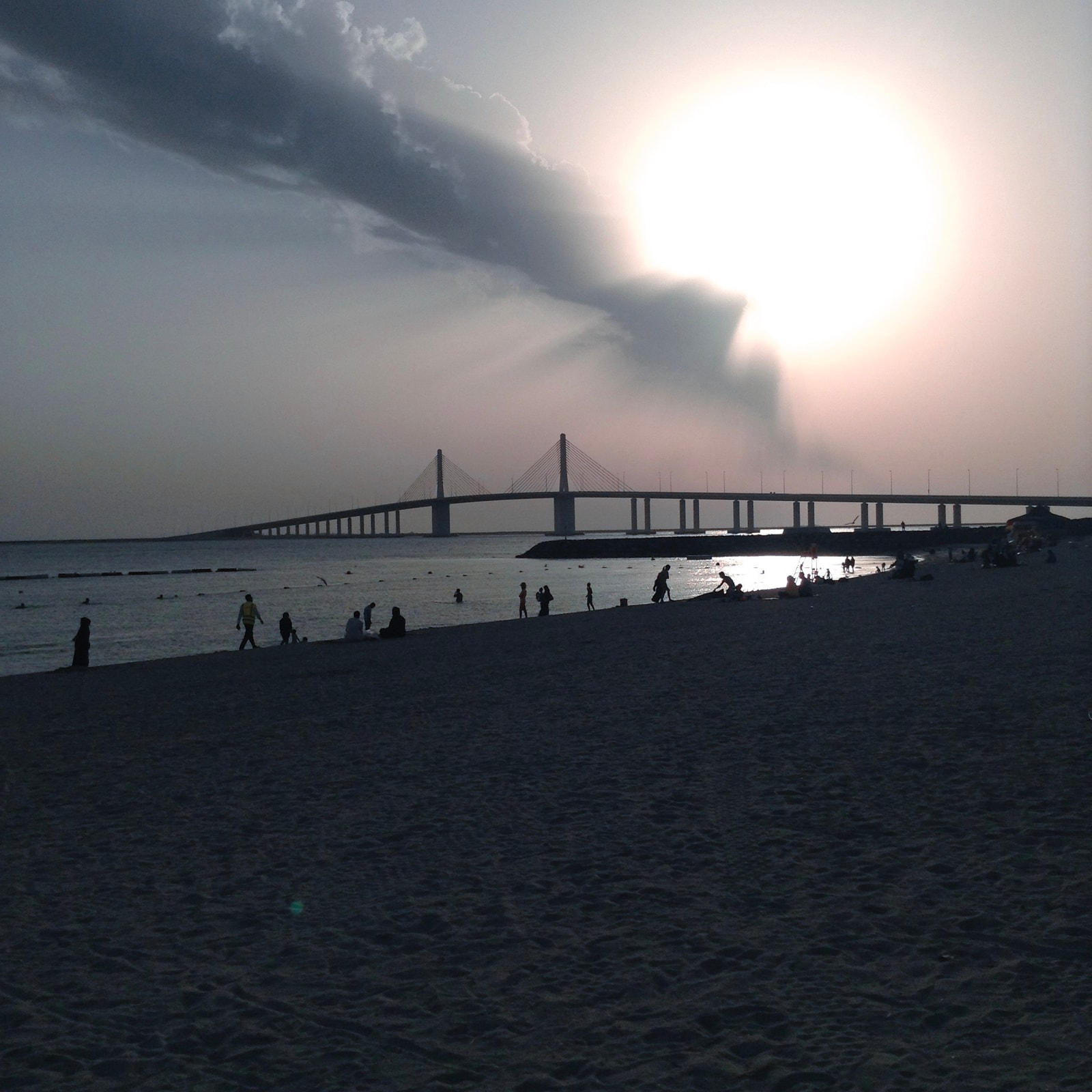 The sun sets behind a bridge at Al Bateen Beach, with dramatic clouds reflected in the calm water and silhouettes of people on the shore.