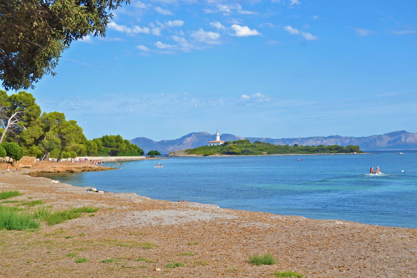 A pebbly beach in the foreground gives way to calm blue water, with a small, green island featuring a lighthouse in the distance.