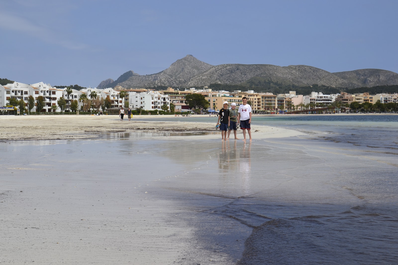 A wide, sandy beach with wet sand reflecting the sky, leading to calm, shallow blue water with distant mountains across the bay.