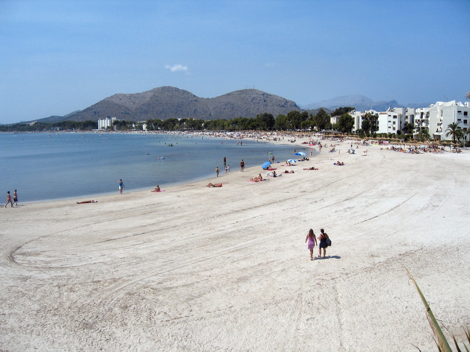 An elevated view from a harbor shows a long, busy sandy beach curving along the coast, with resort buildings and mountains behind.