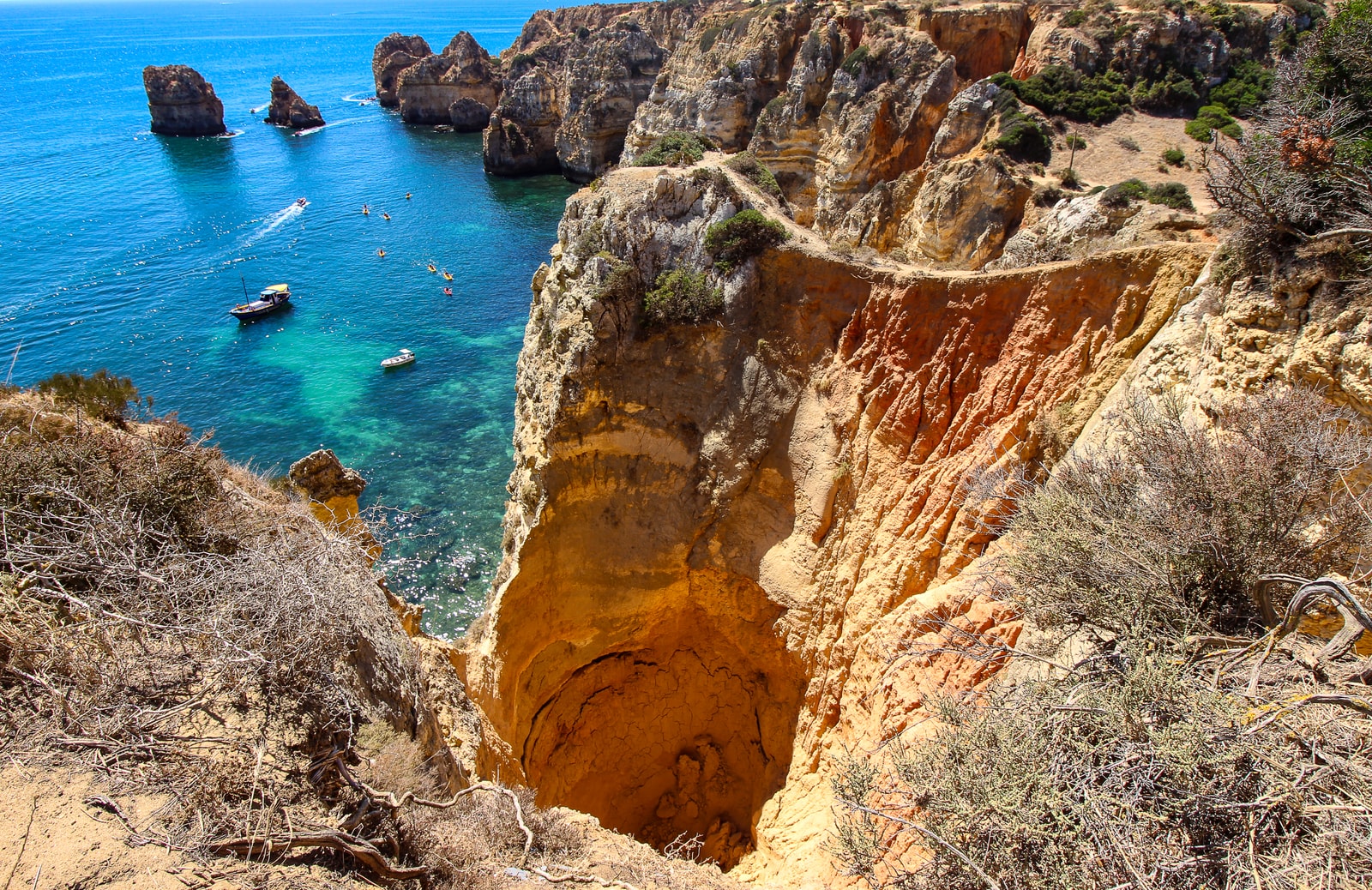A dramatic orange ravine cuts through cliffs to meet a turquoise sea where small boats and swimmers are visible.