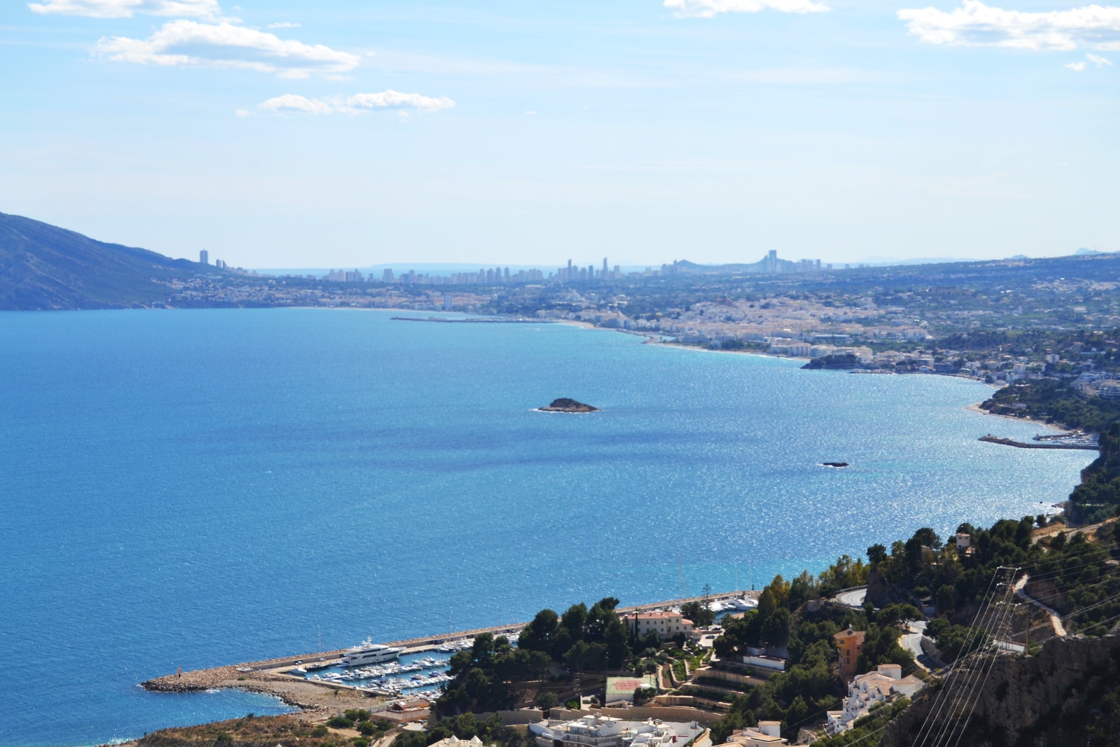 An elevated panoramic view of Altea Bay, showing the small l'Olla island offshore and the distant skyline of Benidorm on the horizon.