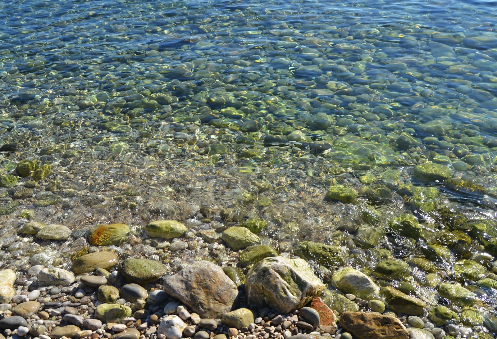 A close-up view of the water's edge at Mascarat beach, showing crystal clear water flowing over smooth, multi-colored pebbles.