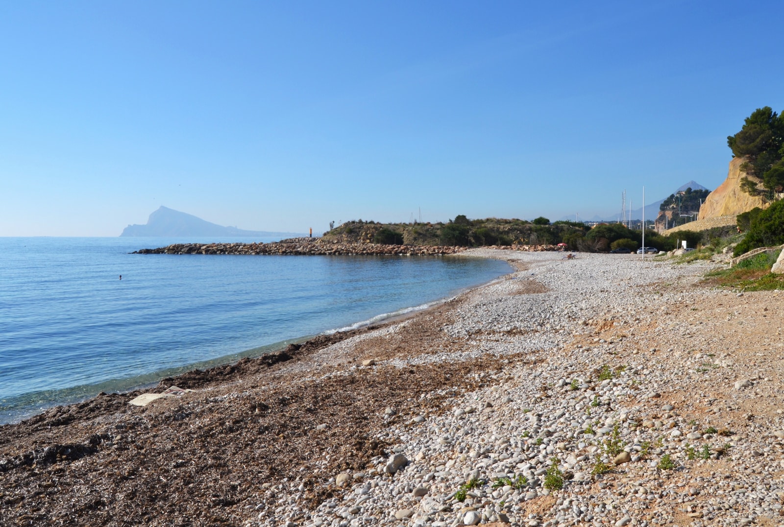 A wide view of the sweeping pebble shoreline at Mascarat beach in Altea, with calm blue water meeting the stones.
