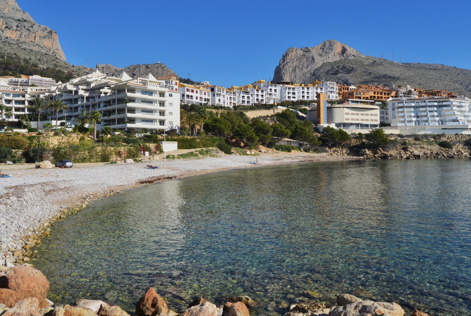 A view of the pebbly Mascarat beach and its bay, with buildings nestled into the surrounding green hills under a bright sky.