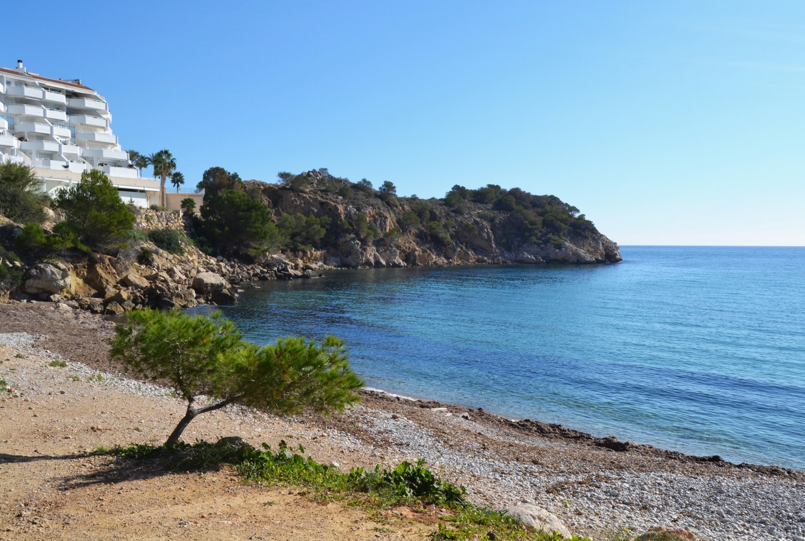 A small pebble beach cove at Mascarat, framed by a rocky point with a windswept tree in the foreground.