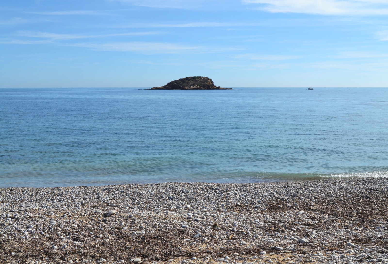 A tranquil view of Playa de l'Olla's pebble beach with its small, rocky offshore island under a blue sky.