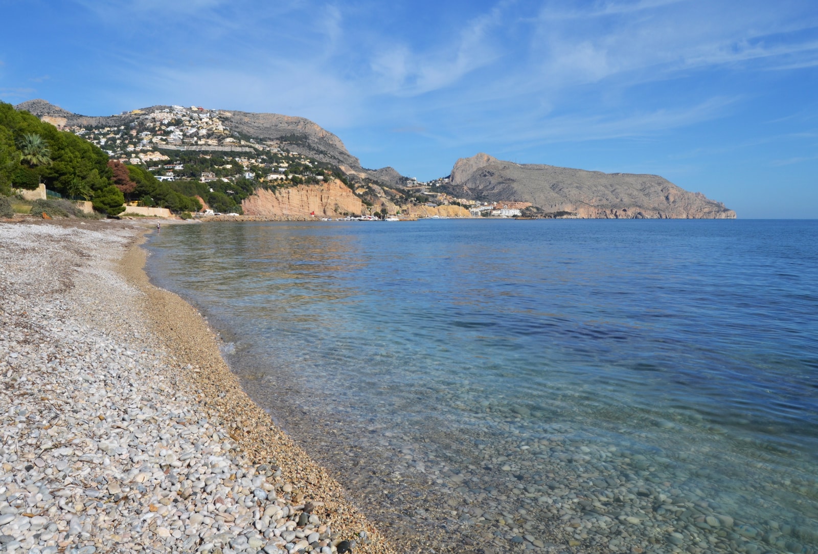 A curving pebble beach at Playa de l'Olla in Altea, with exceptionally clear, turquoise water in the shallows revealing the smooth stones below.