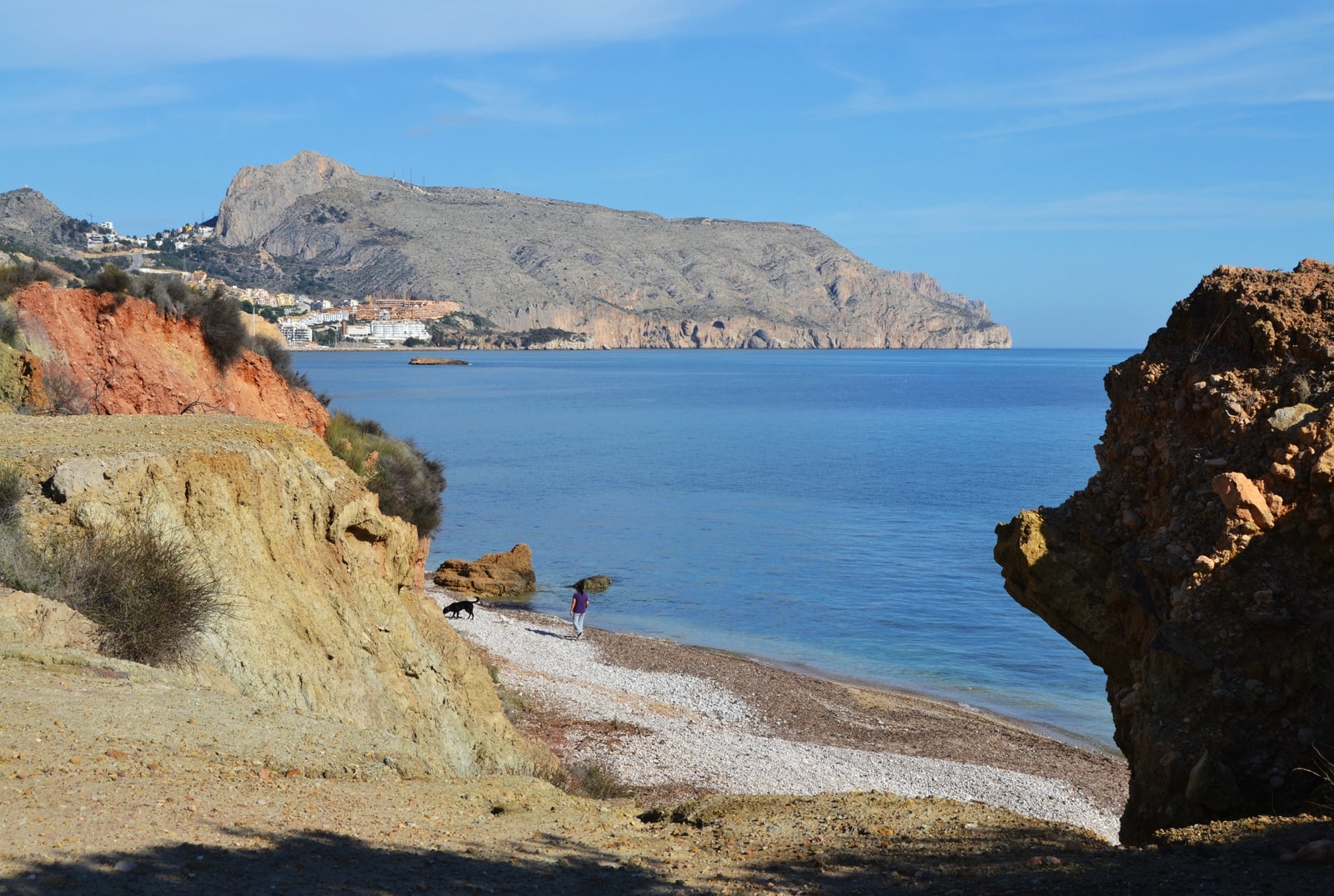 The pebbly shore of Playa de l'Olla in Altea meets the calm blue sea, with the rugged Morro de Toix headland in the background.