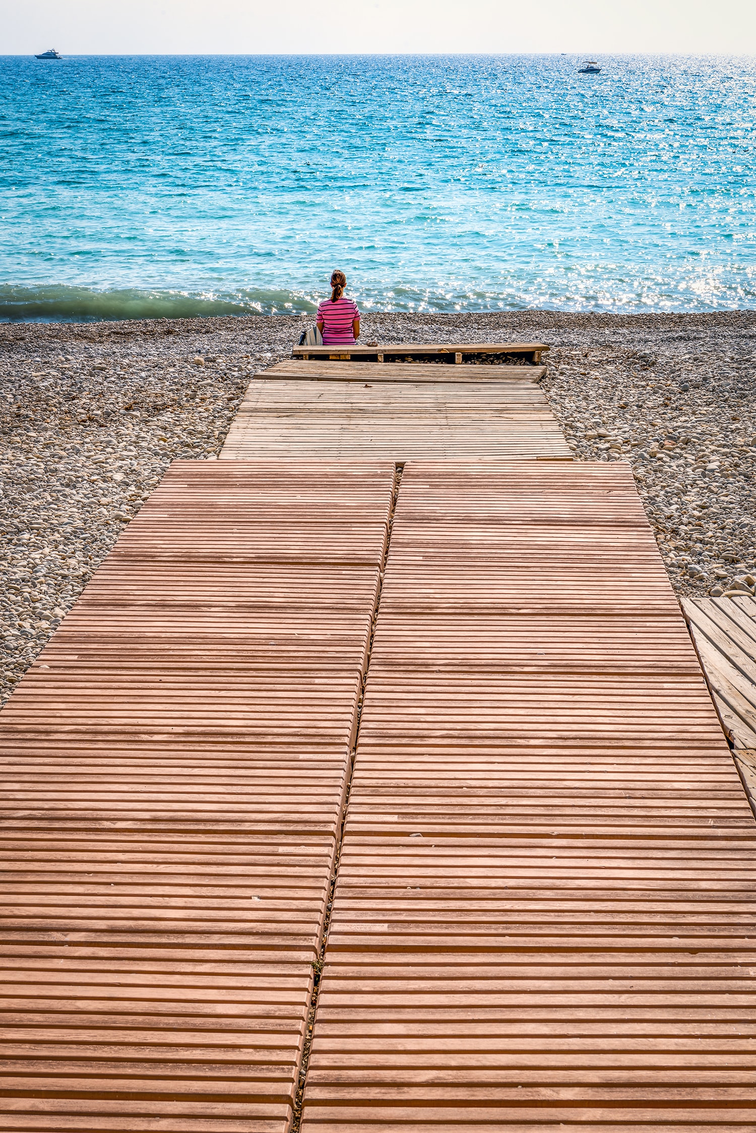 A wooden boardwalk leads across a wide pebble beach towards a calm blue sea, with a lone figure in the distance.