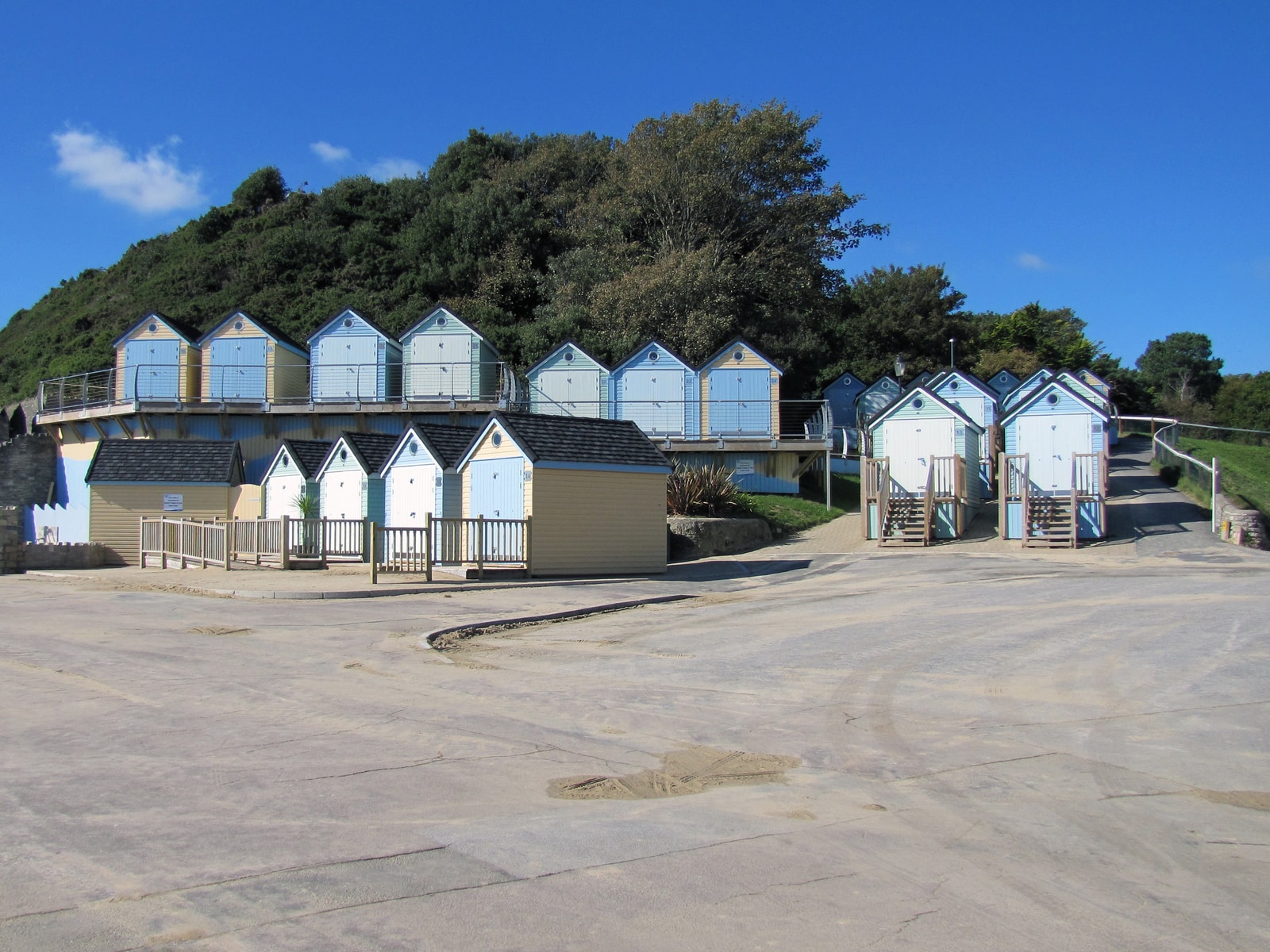 Pastel-colored beach huts are lined up at Alum Chine, with a lush wooded hill in the background.