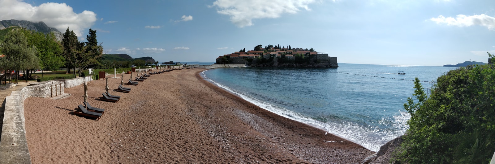 A wide panoramic shot shows a pink-sanded beach lined with neat rows of loungers and umbrellas, leading towards the fortified island resort of Sveti Stefan.