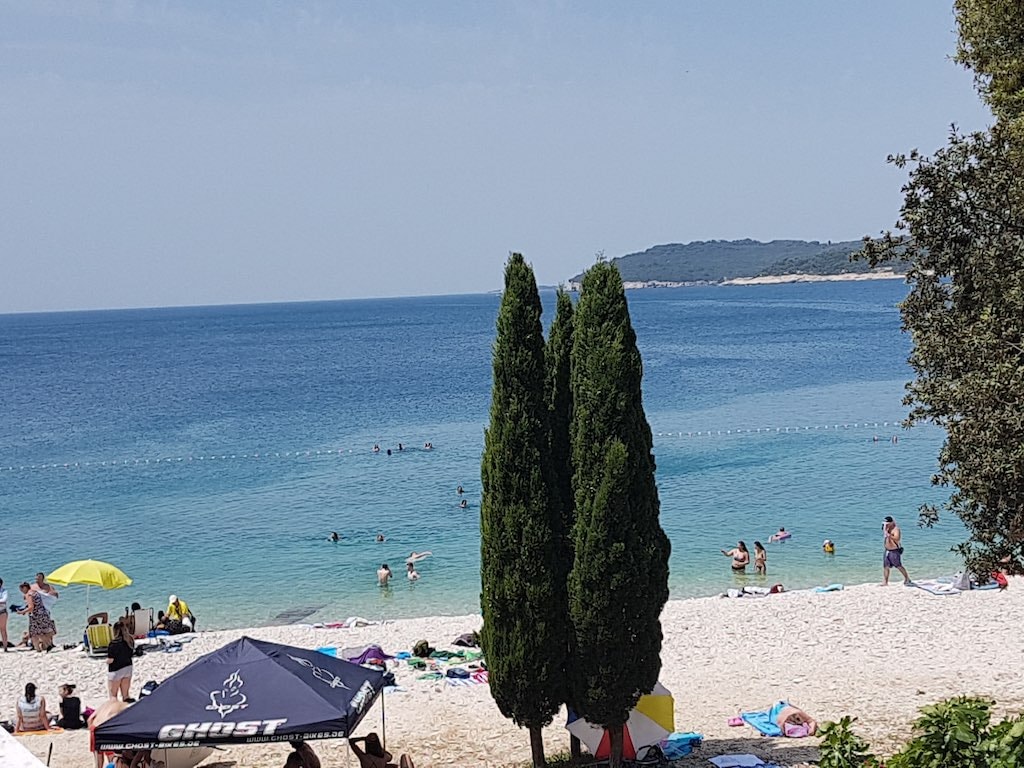 Sunbathers relax on the white pebbles of Ambrela Beach, framed by tall, dark green cypress trees and the turquoise Adriatic Sea.