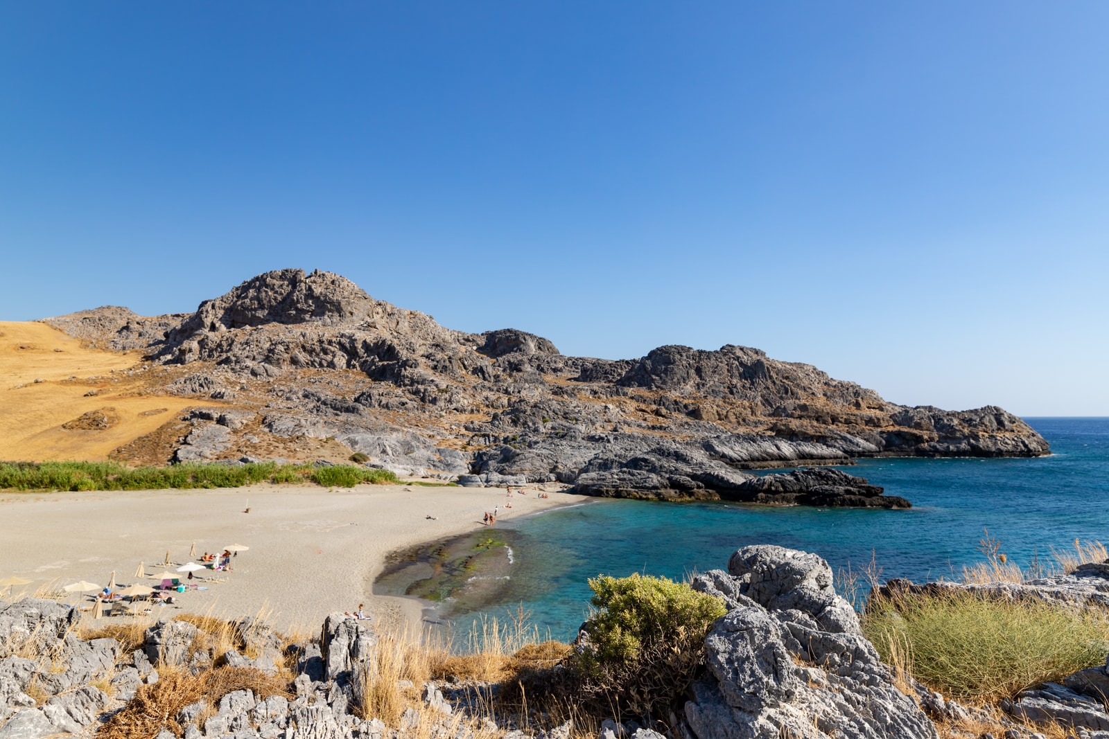 An aerial view of a sheltered, curved sandy bay with turquoise water and umbrellas on the shore.