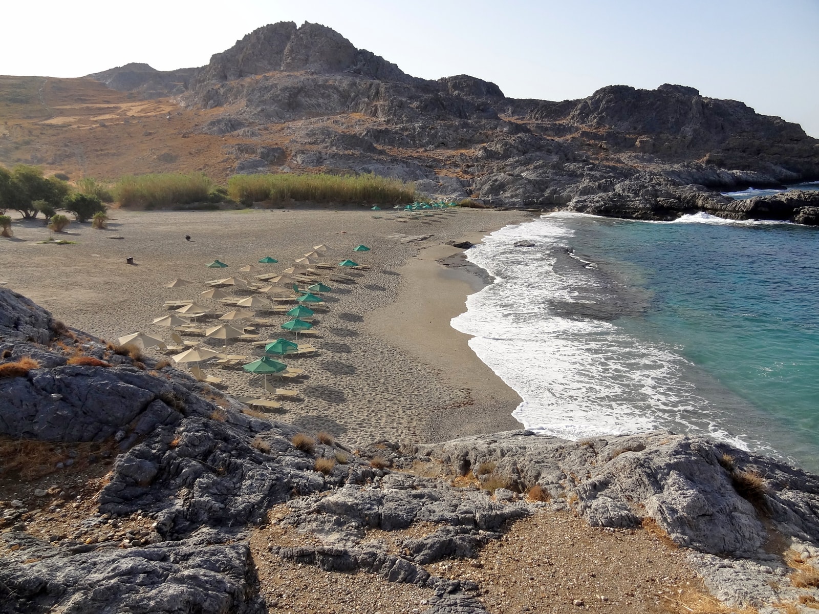 An elevated view of a pebbly beach in a cove with turquoise water and dark, rocky hillsides.