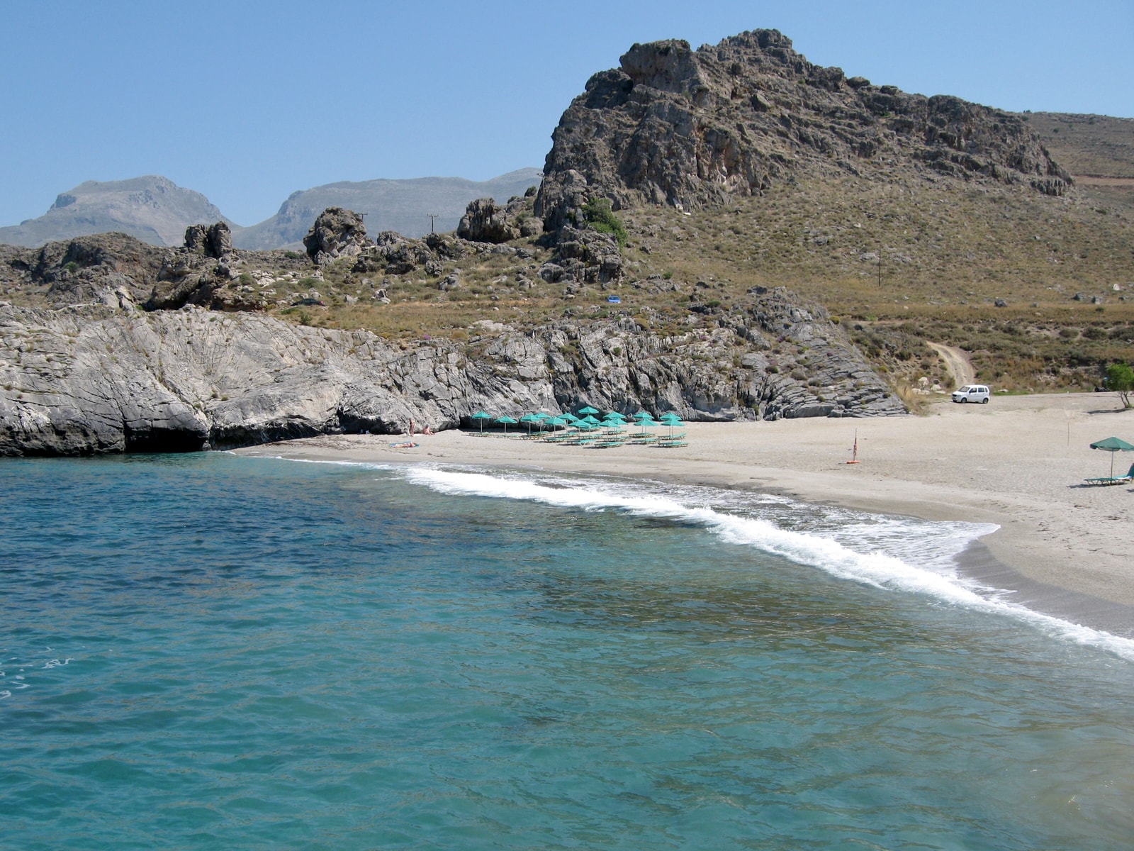 A view of a crescent-shaped beach with turquoise water and rocky outcrops, backed by dry mountains.