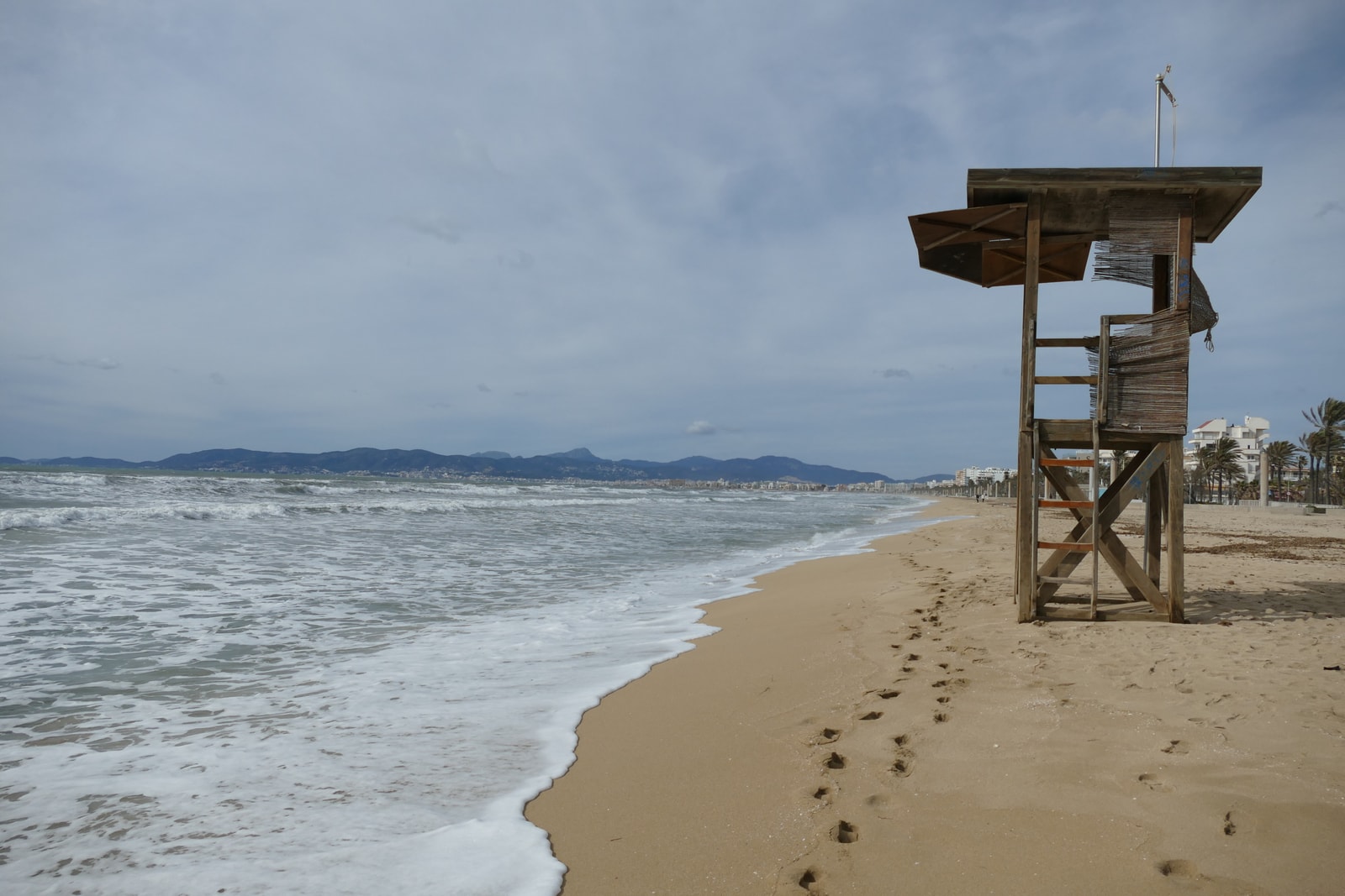 A lifeguard tower stands on a vast, empty sandy beach, with footprints in the sand and resort buildings visible in the distance.