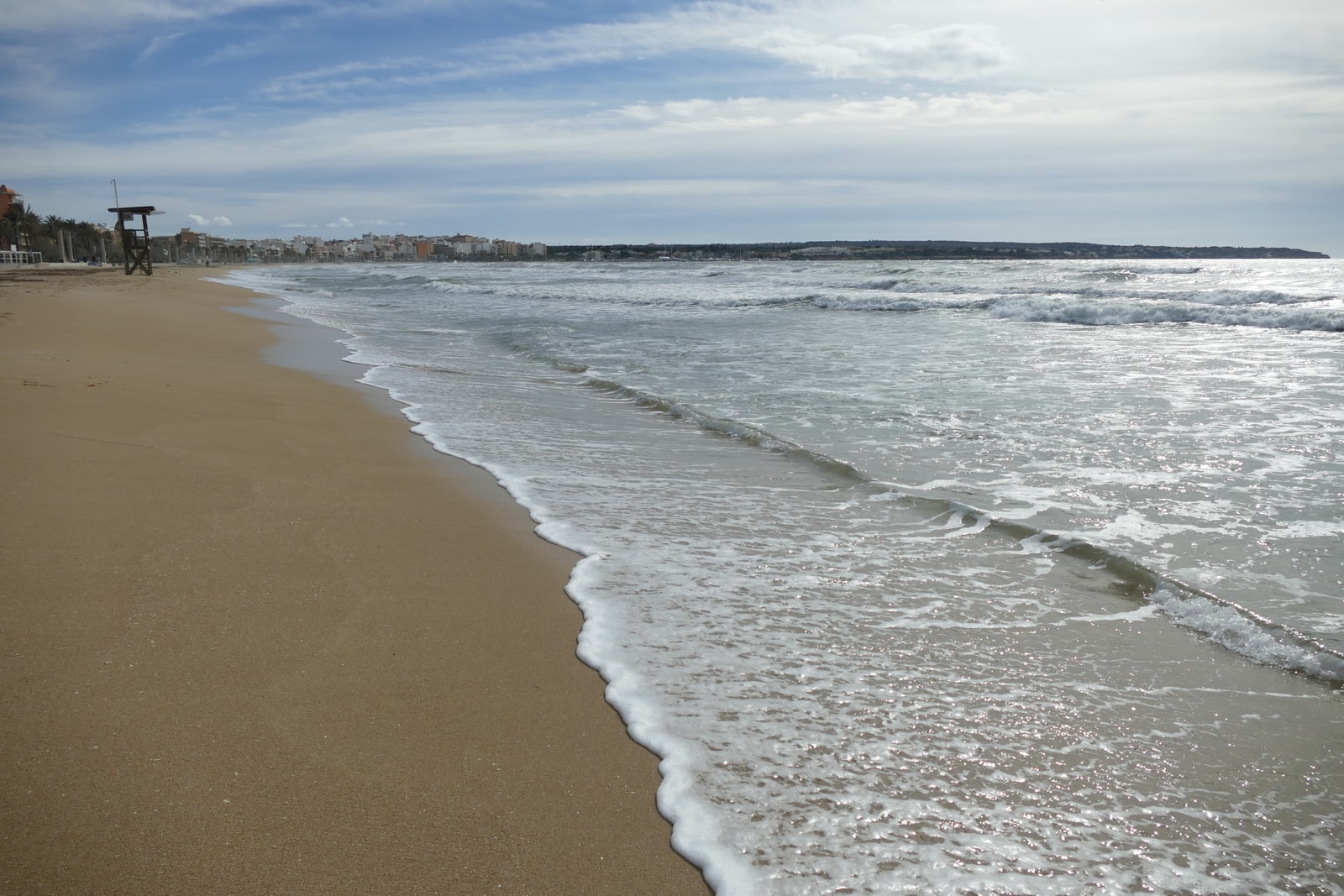 Sunlit waves lap against the sandy shore at Playa de Palma, with a lifeguard station in the distance.