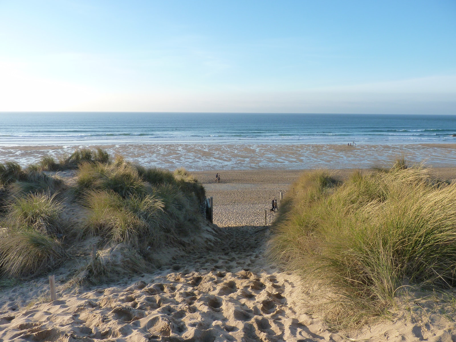 A path leads through grassy sand dunes towards the sea at Fistral Beach.
