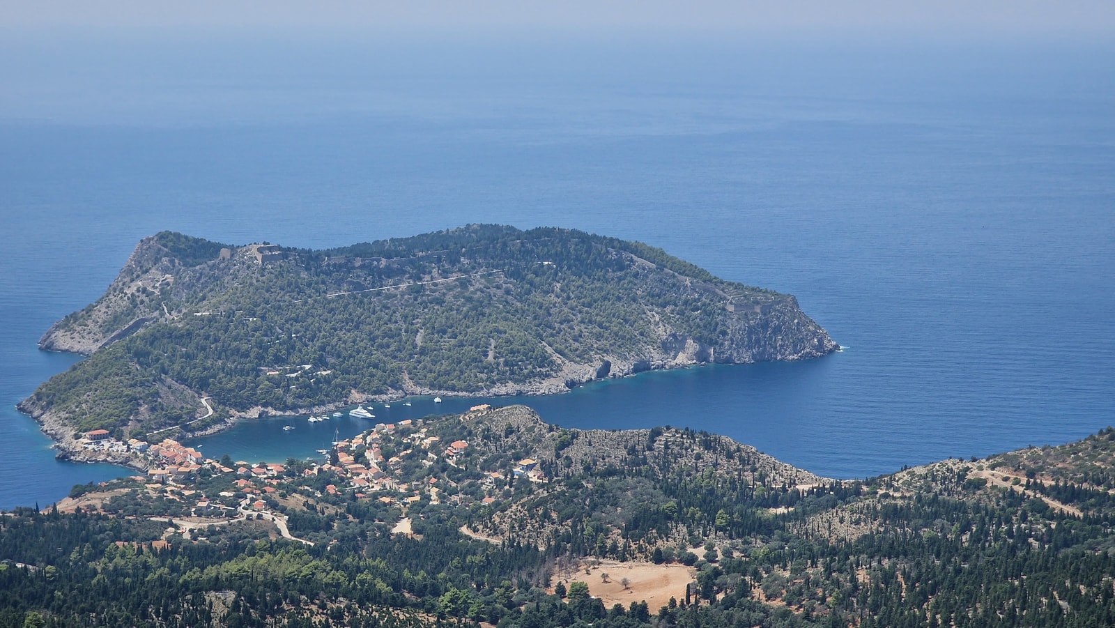 A panoramic view of Assos village nestled on a narrow peninsula, flanked by two bays and sitting below a large, forested hill.