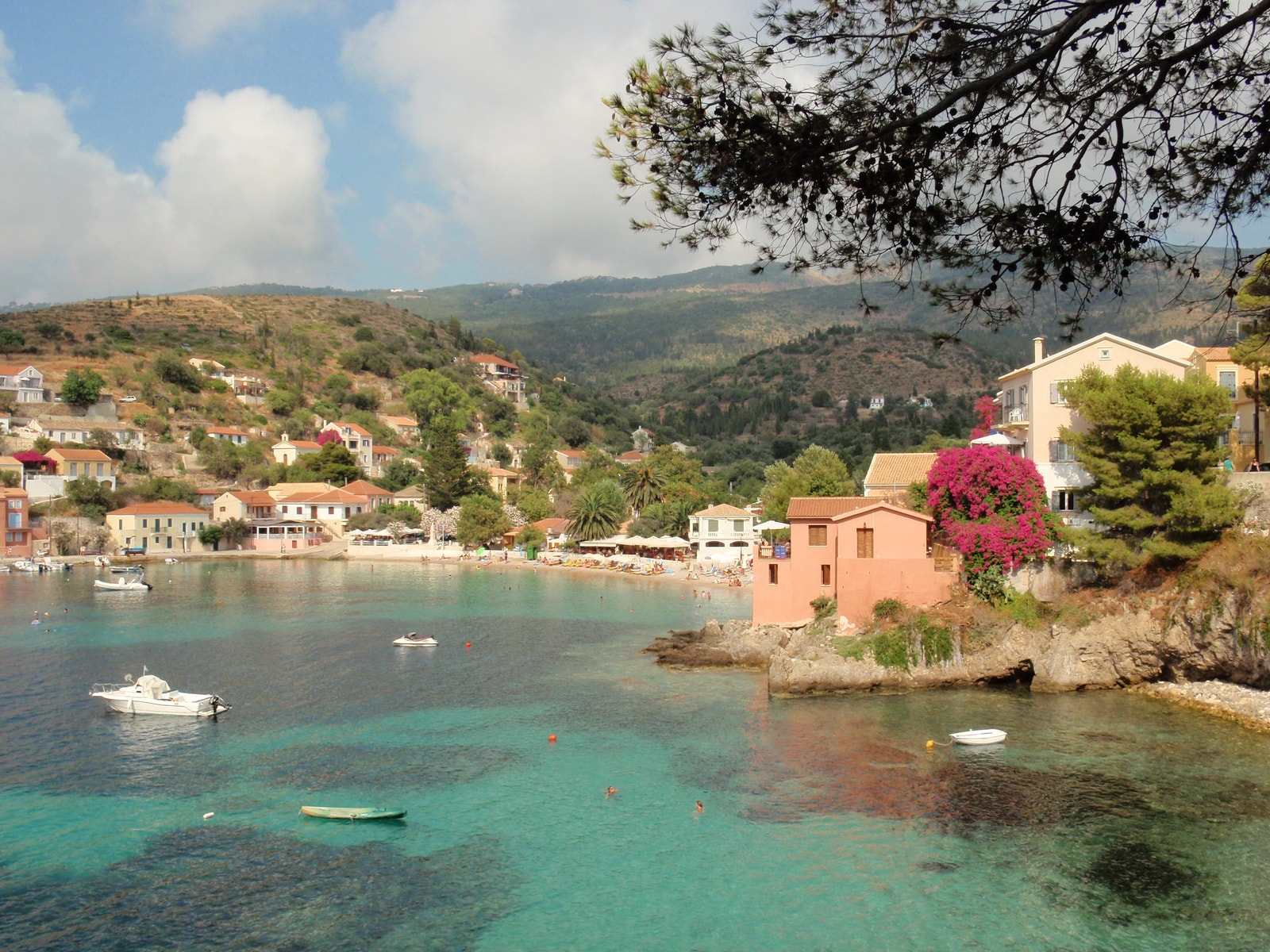 The colorful houses of Assos village with pink bougainvillea, overlooking small boats in a calm turquoise bay.