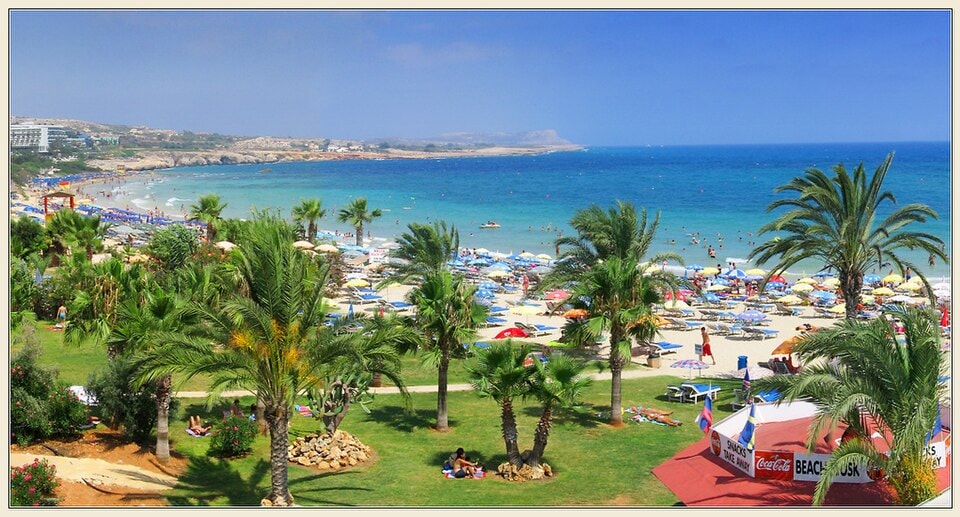 A view of a busy resort beach in Ayia Napa, with rows of sunbeds and umbrellas on the sand next to calm, turquoise water.