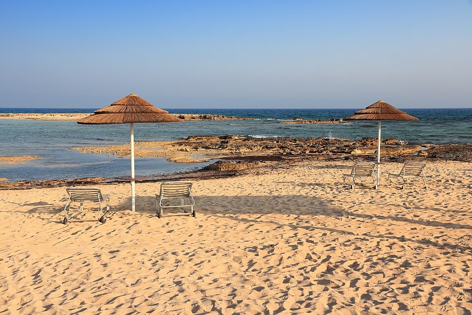 A quiet, sandy beach with rows of empty sun loungers and thatched umbrellas facing calm, shallow water under a bright sky.