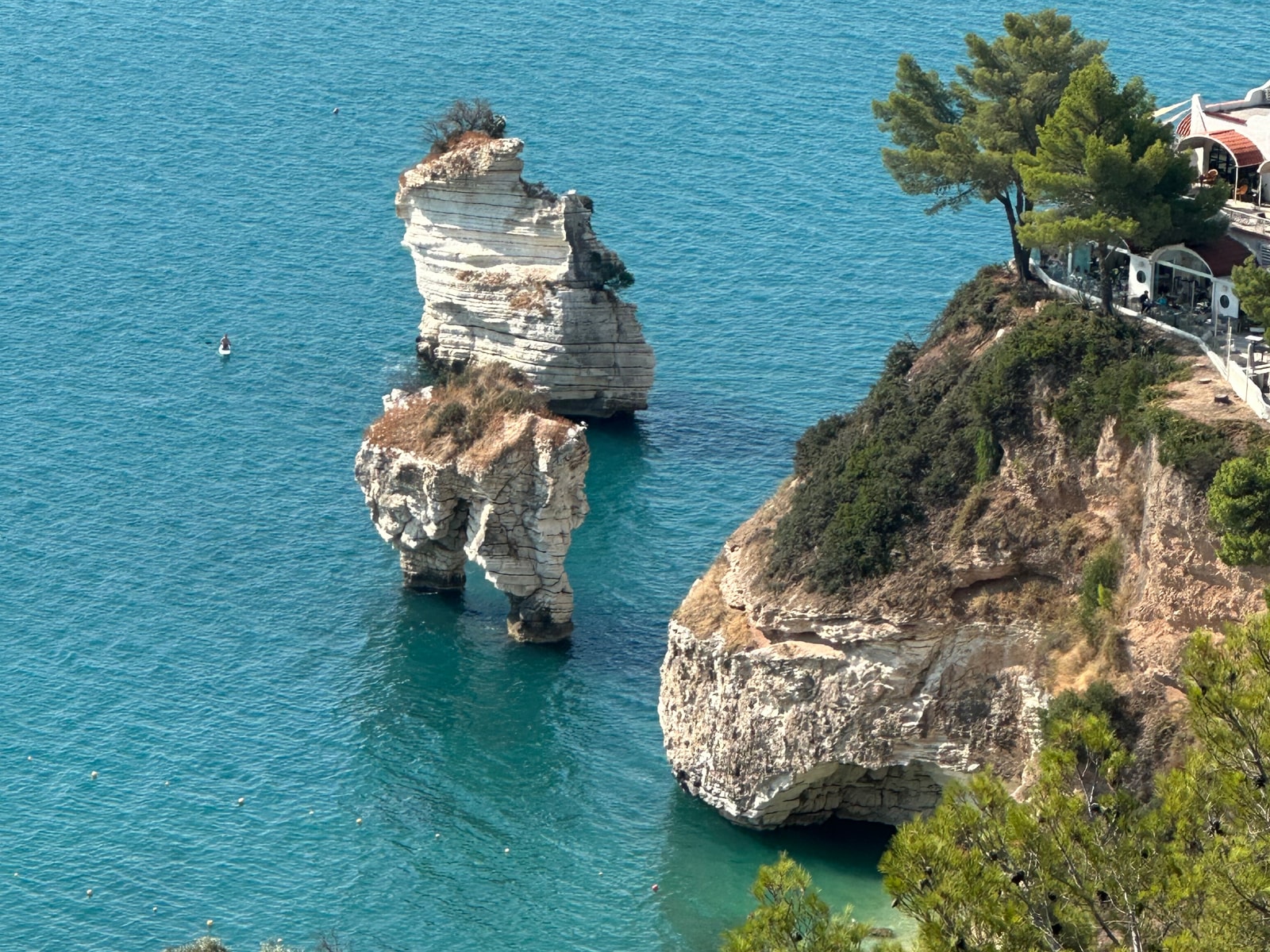 A close-up view of two massive, wave-sculpted white limestone sea stacks rising from the turquoise sea at Baia delle Zagare.