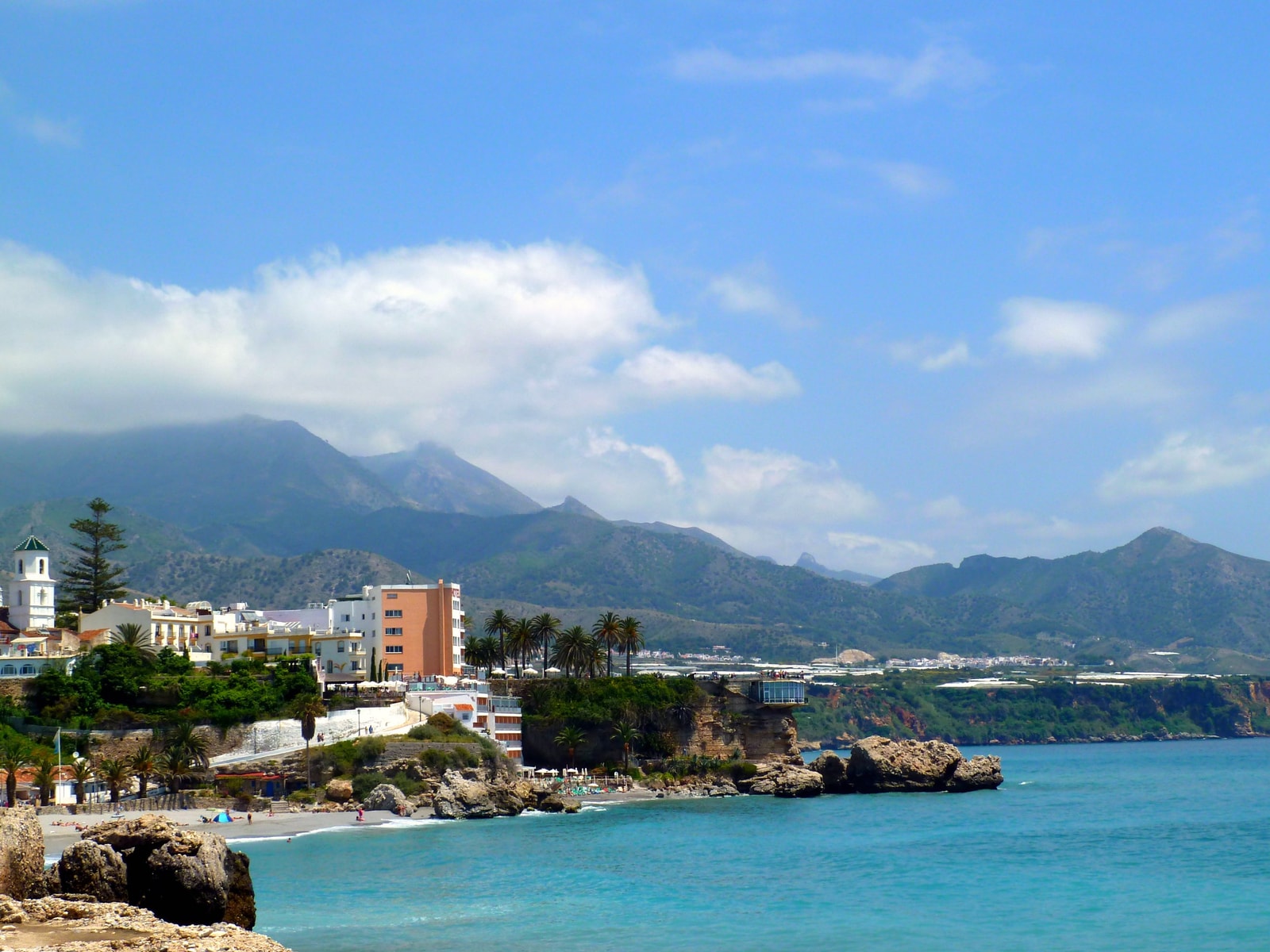 An elevated view of Nerja's Balcón de Europa shows the promenade jutting out into the blue sea, with small beaches nestled into the cliffs on either side.