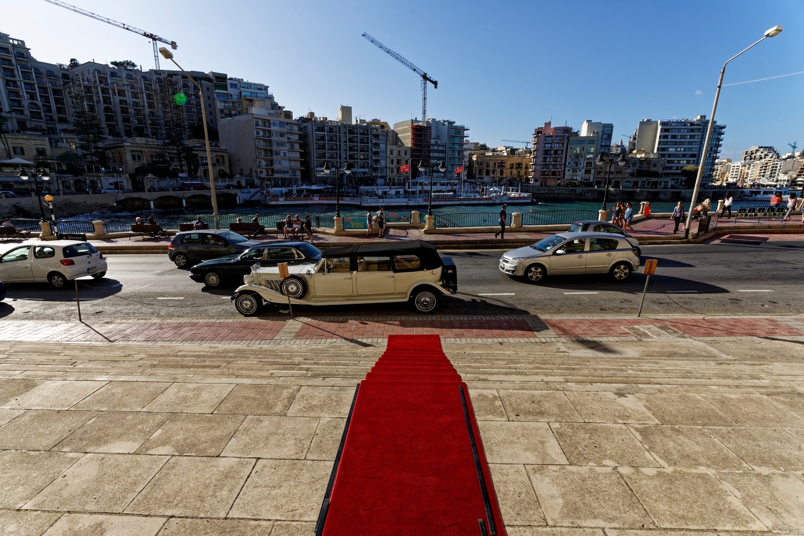 A classic luxury wedding car parked on the street near the stone steps leading down to Balluta Bay, with the parish church visible.