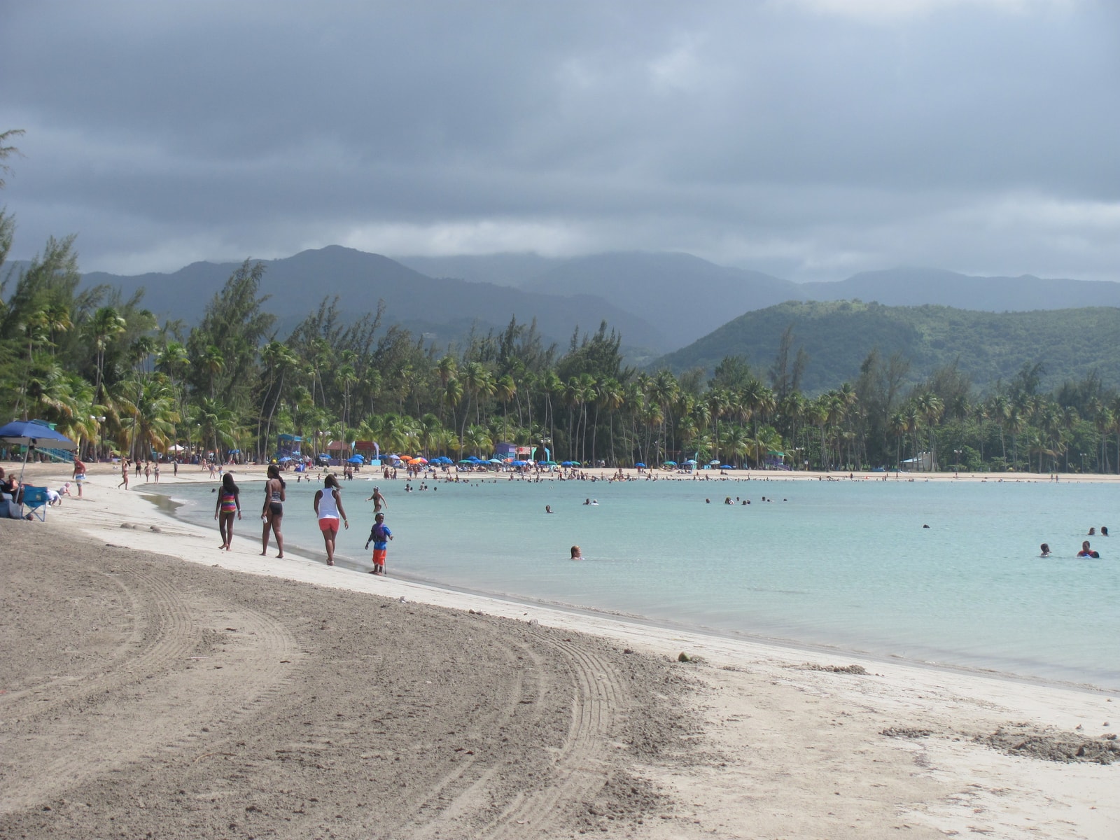 A broad view of the public beach at Luquillo, showing the sandy shore, palm trees, and mountains in the hazy distance.