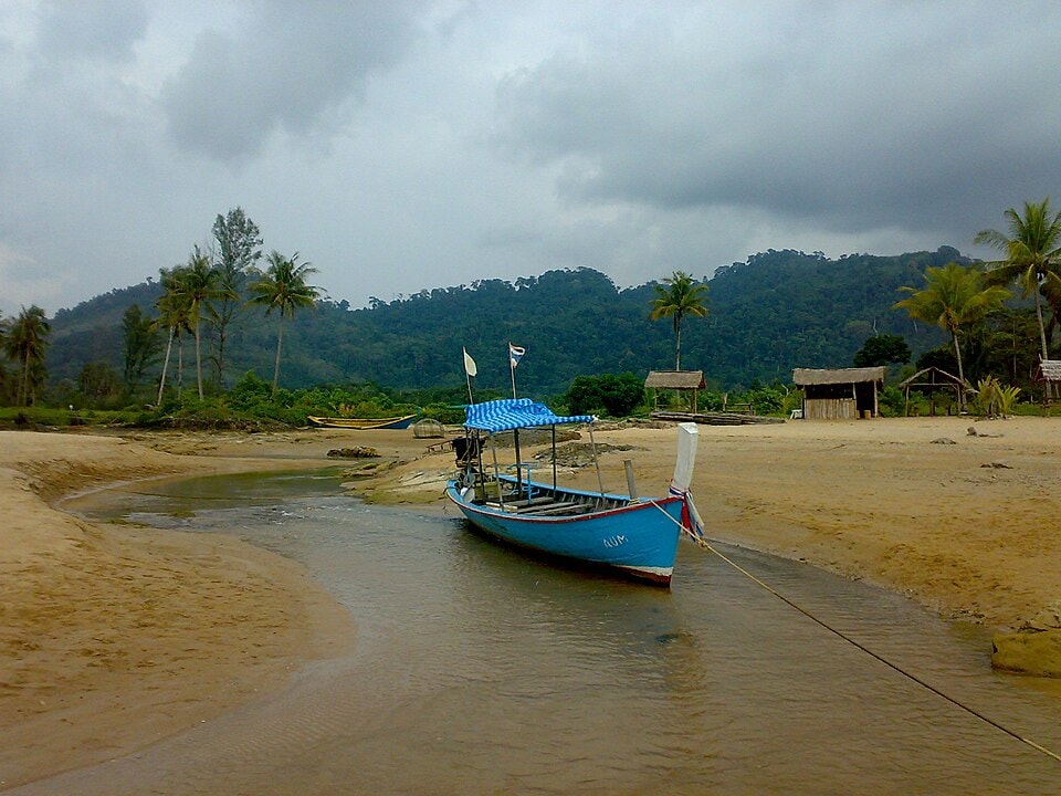 A colorful long-tail boat rests in a tidal channel at Bang Niang Beach, with forested hills in the background under a cloudy sky.