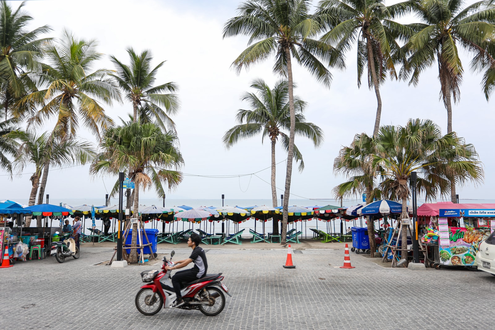 A view of Bang Saen's palm-lined promenade, with beach umbrellas, food stalls, and the sea in the background.