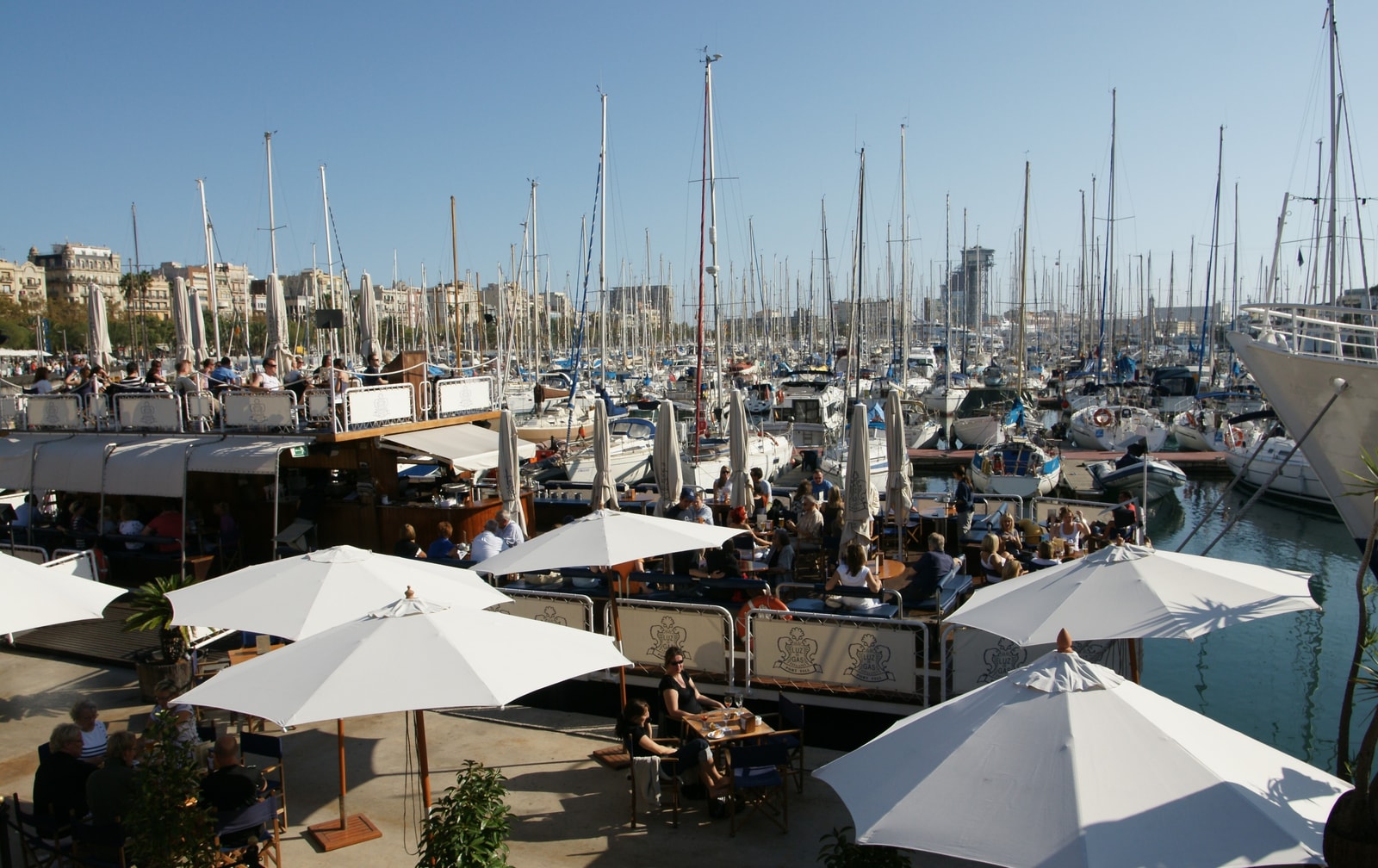 A view of the Port Vell marina with docked sailboats and waterfront cafes under a clear blue sky.