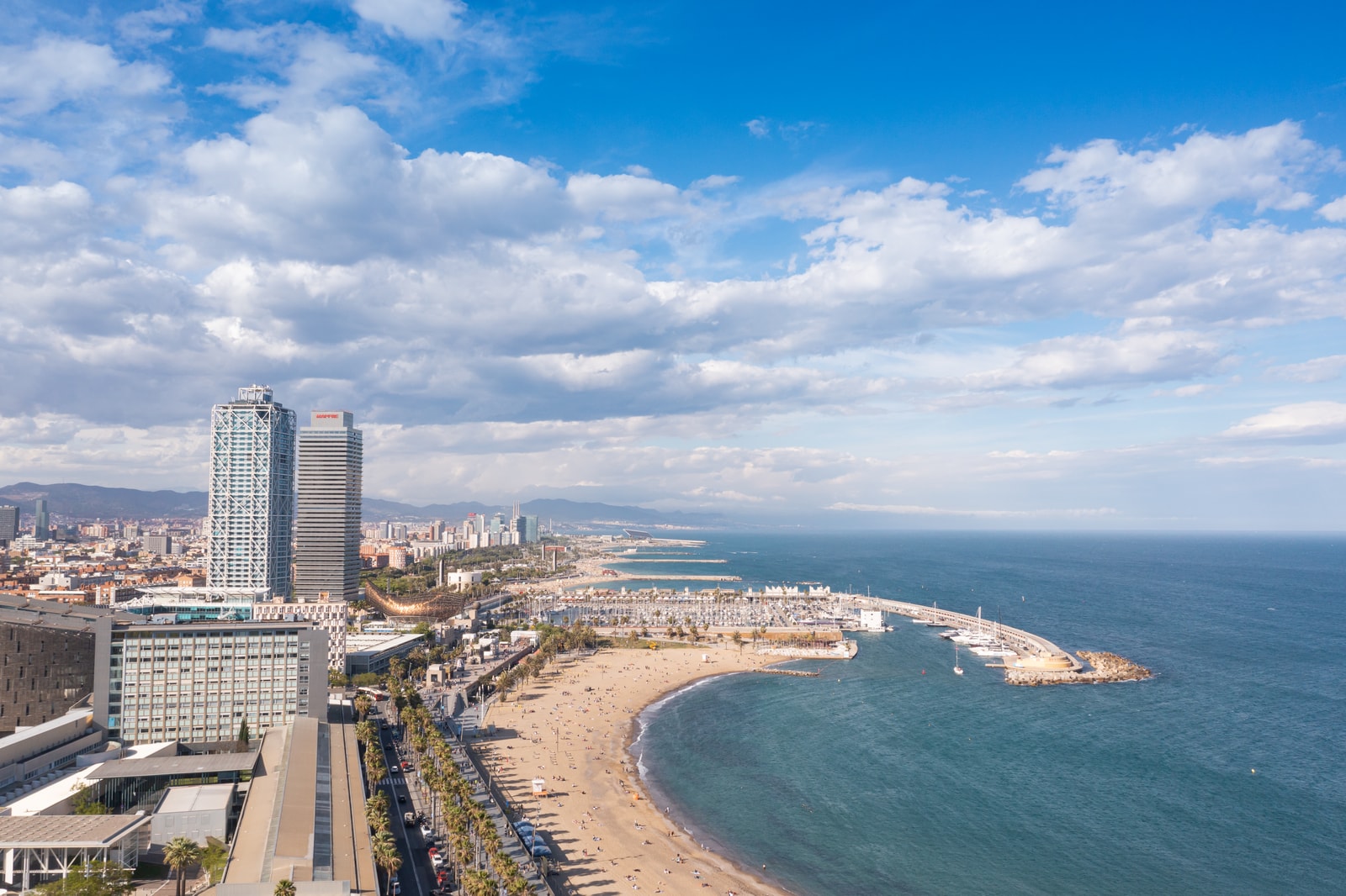 A wide aerial view shows Somorrostro Beach, the Port Olímpic marina, and the twin skyscraper towers in the background.
