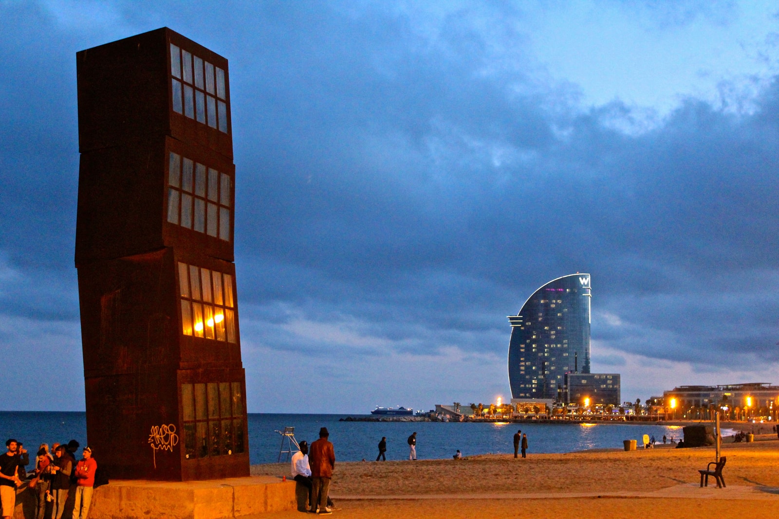 A sculpture of four stacked, rusted metal cubes stands on the beach against a dramatic sunset sky.