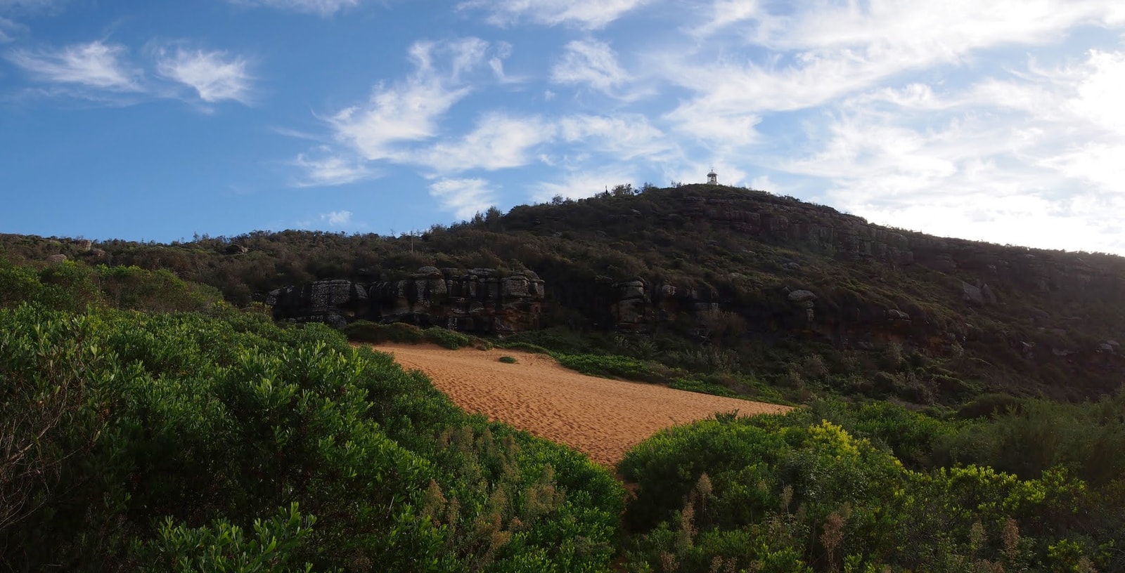 A view of the rocky, vegetated Barrenjoey Headland under a blue sky, illustrating a key part of Sydney's coastal walk scenery.