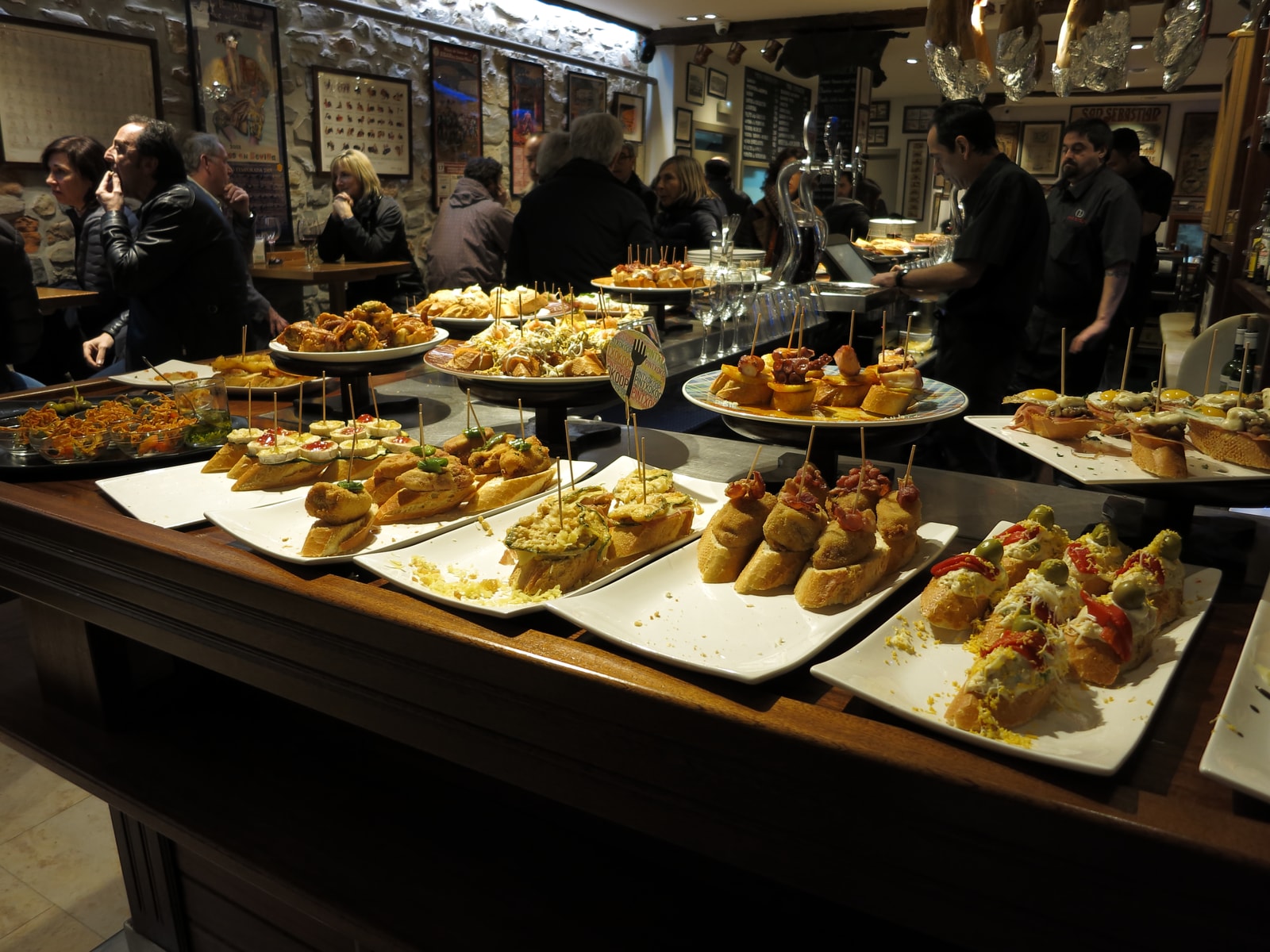 An inviting display of various pintxos on a wooden bar top in a bustling San Sebastián tavern.