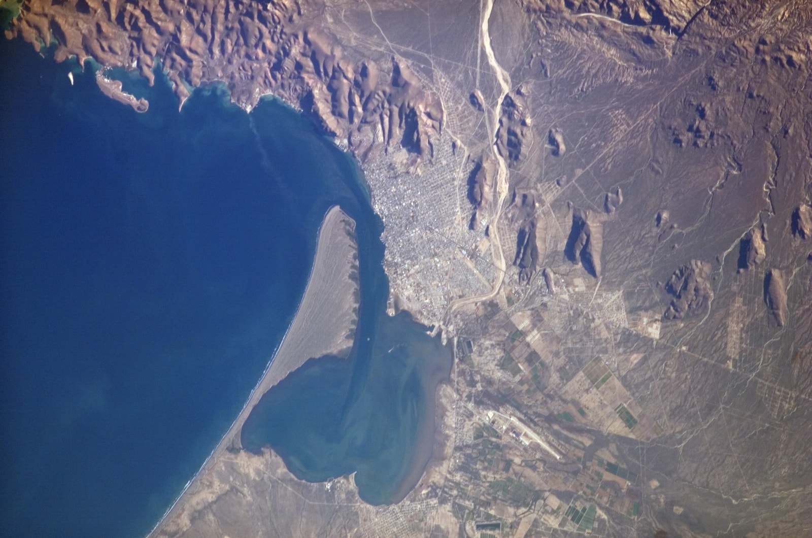 An aerial photograph of the Bay of La Paz, showing the city's grid layout meeting the water, with a long desert peninsula in the background.